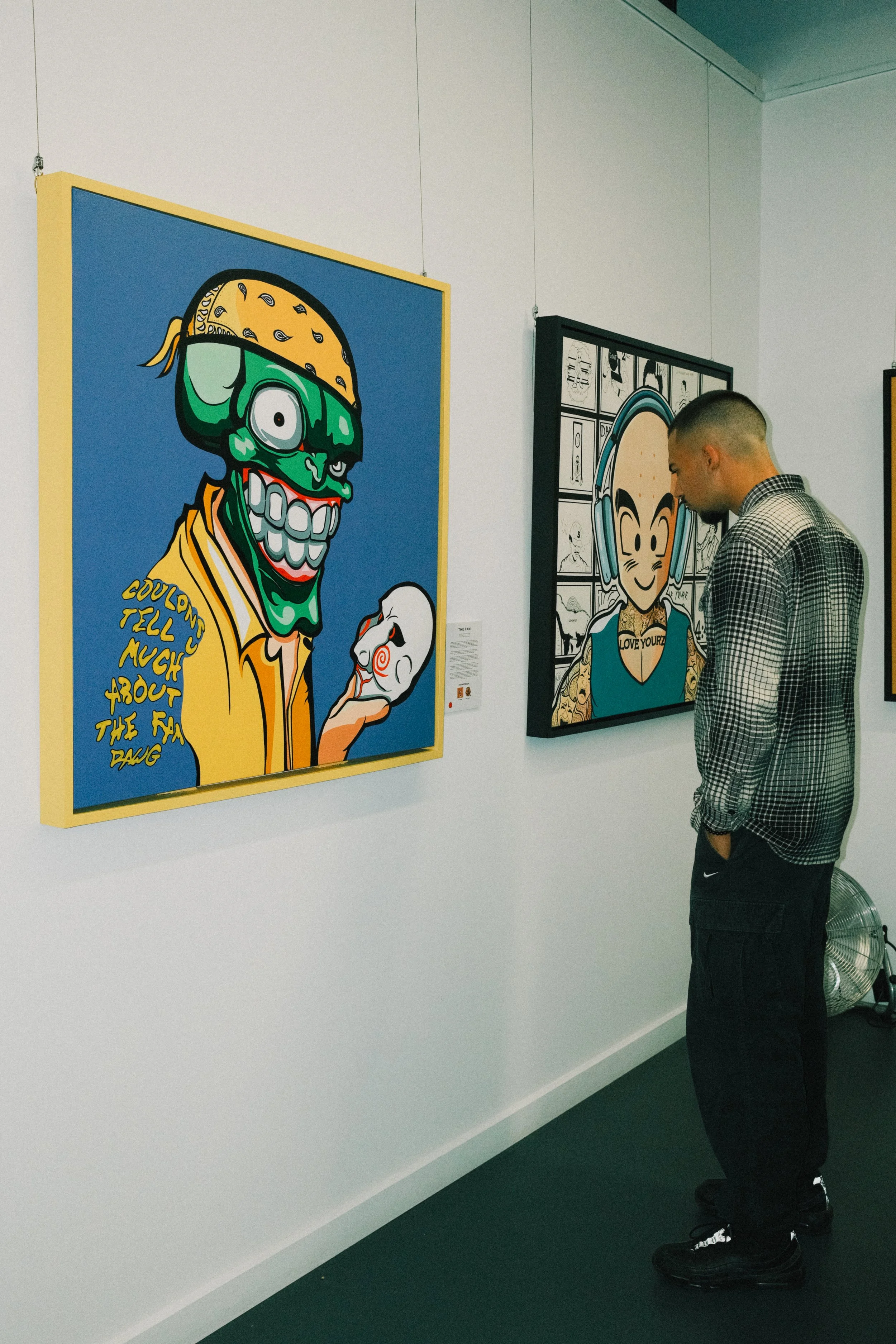A man in a checkered shirt observes colorful cartoon artwork on display in an art gallery. The artwork includes a green, one-eyed, zombie-like character holding a mask, with text that reads "Couldn’t tell much about the fad dawg."
