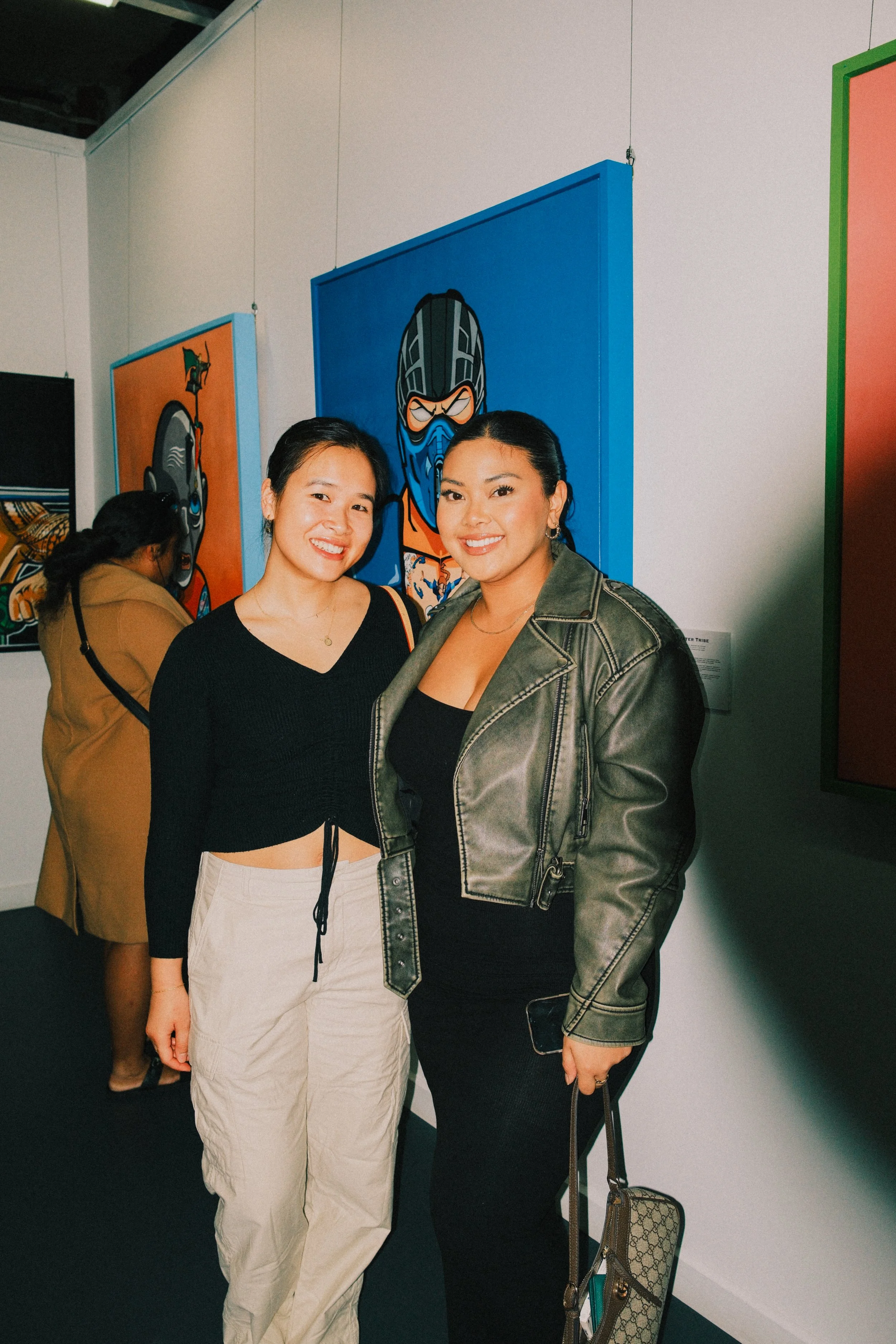 Two women posing for a photo at an art gallery, with colorful paintings in the background. One woman is wearing a black crop top and beige pants, and the other is wearing a black top, green jacket, and carrying a designer purse.
