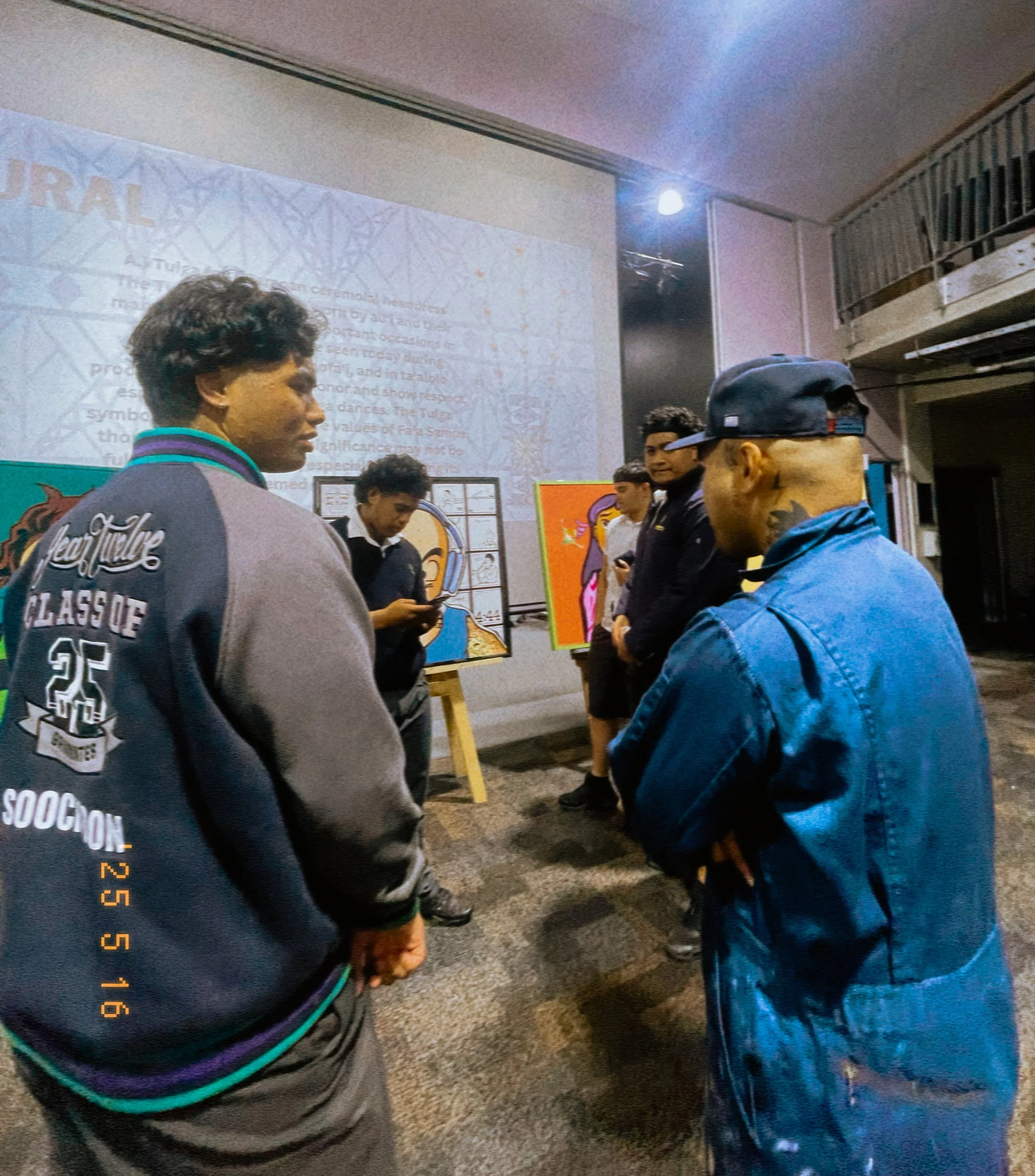 A group of young people standing in a circle indoors, possibly at an art exhibit or workshop, with colorful artwork displayed behind them.
