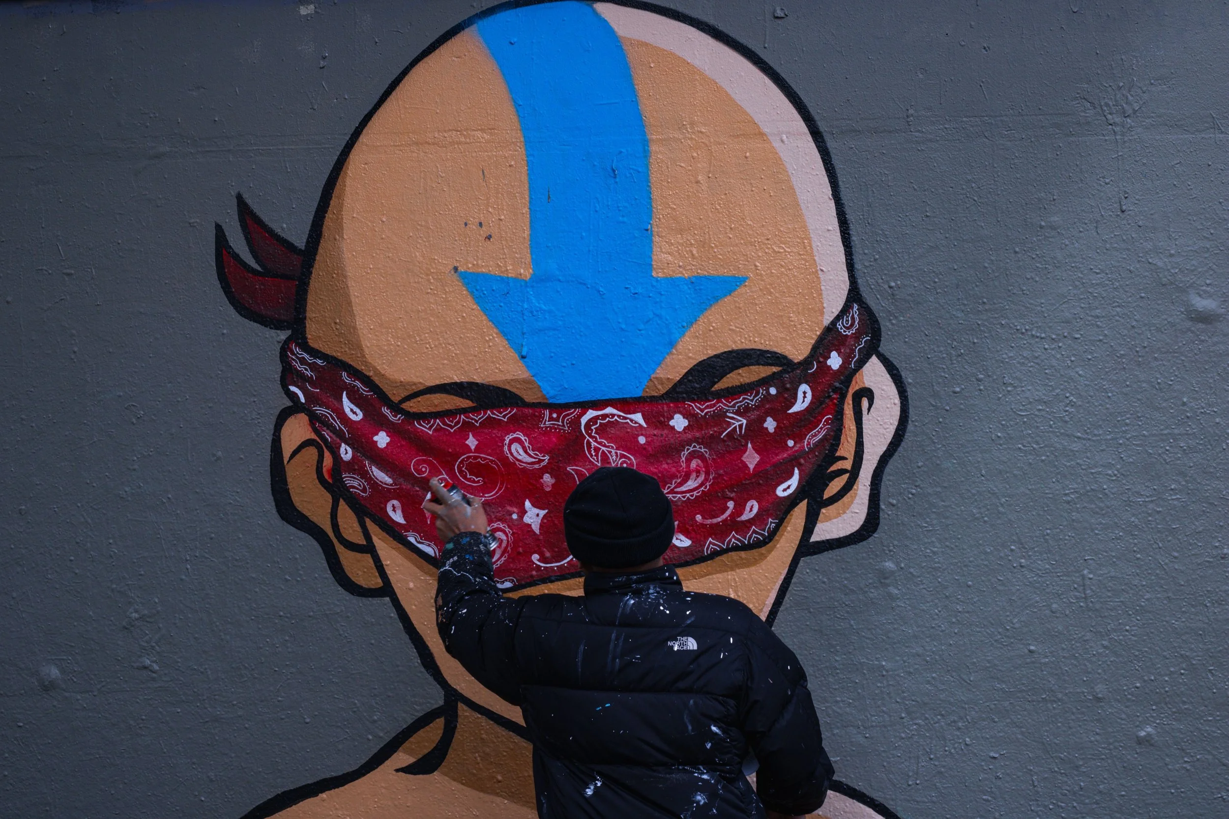 An artist spray-paints a mural of a person with a red bandana covering their eyes on a gray wall. The person's head is depicted with a large blue arrow pointing down on their forehead.