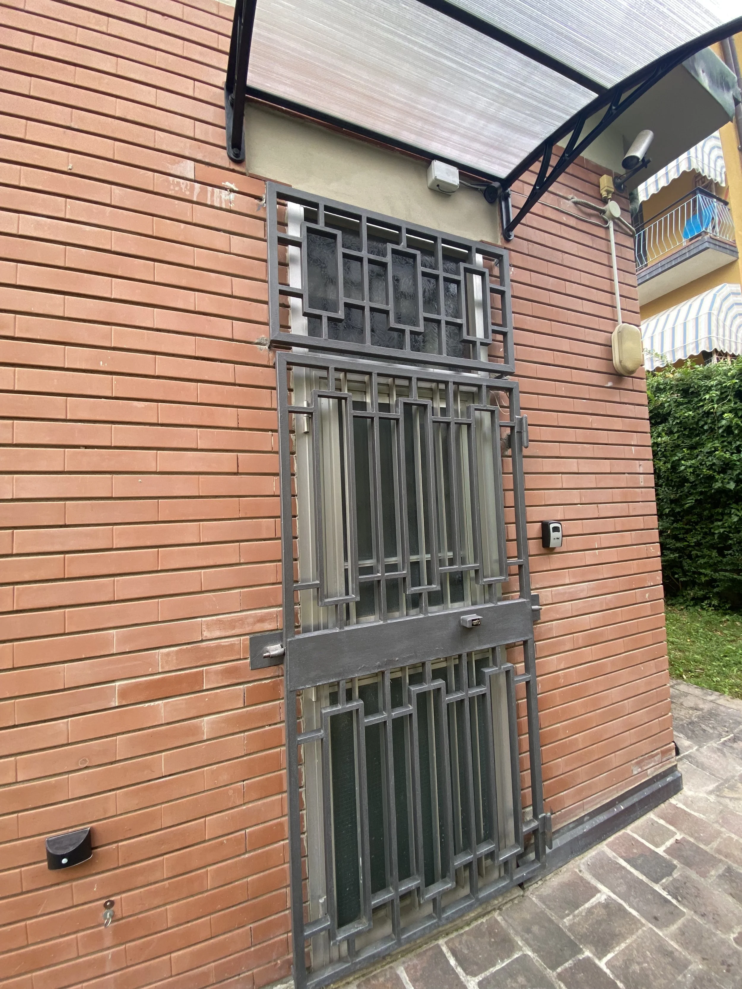 Porta di ferro con sbarre e inferriate sulla facciata di un edificio in mattoni rossi, con tettoia sopra e dettagli come sensori e cavi elettrici.
