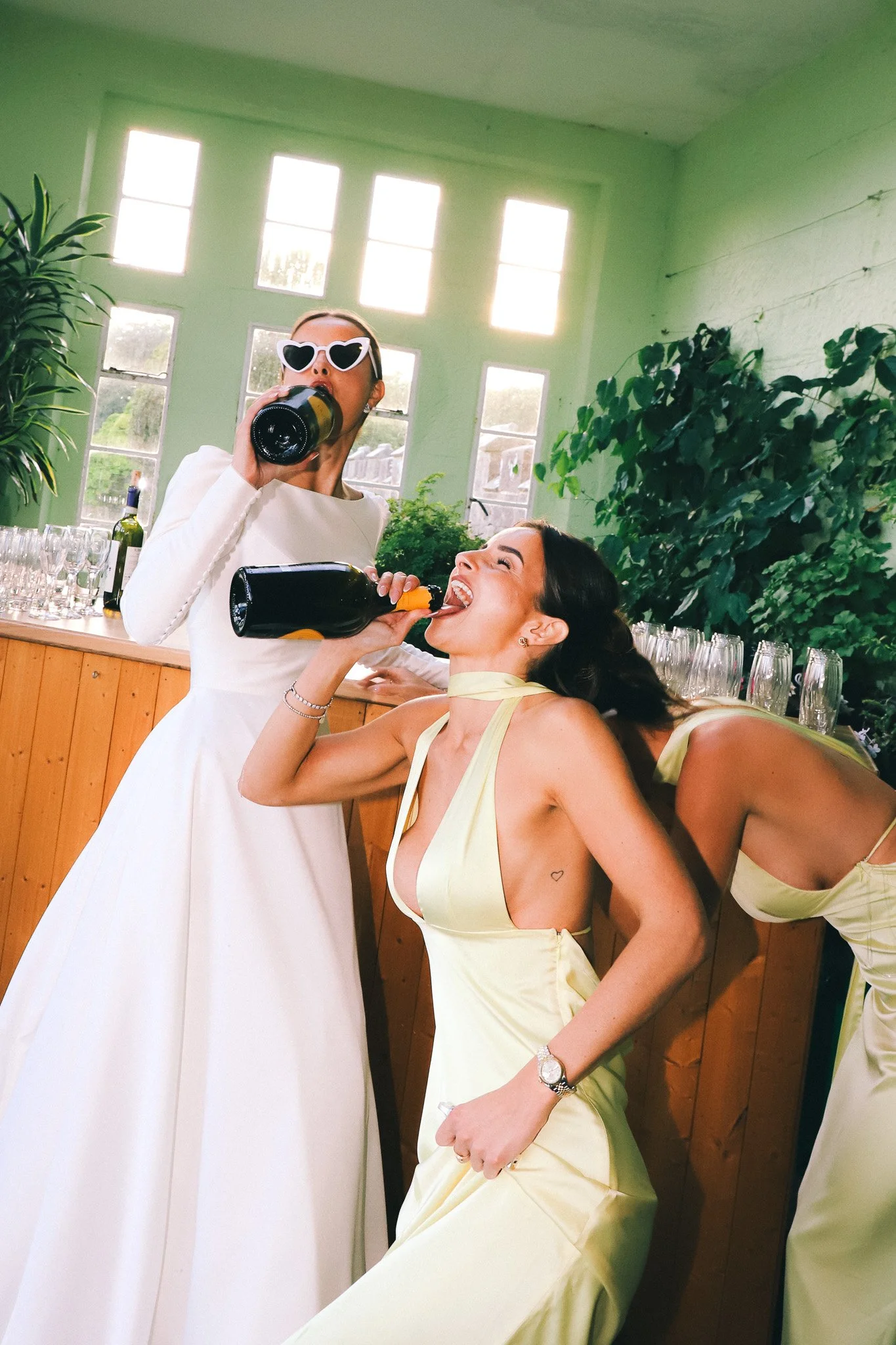 Two women celebrating with drinks at a party; one in a white dress with heart-shaped sunglasses, the other in a yellow dress, drinking from bottles, with glasses and bottles on the counter behind them.