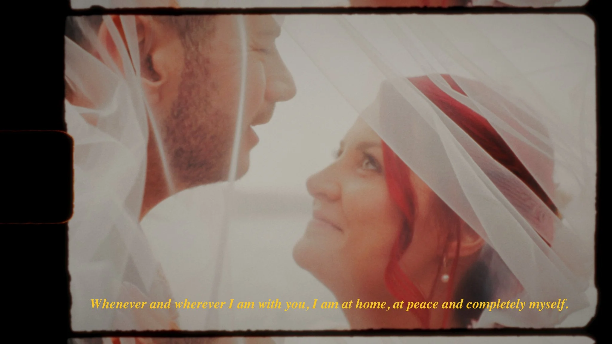 A close-up of a man and a woman, both smiling, with the woman wearing a white veil with red accents, on their wedding day. The image has a quote in yellow text at the bottom that reads, "Whenever and wherever I am with you, I am at home, at peace and