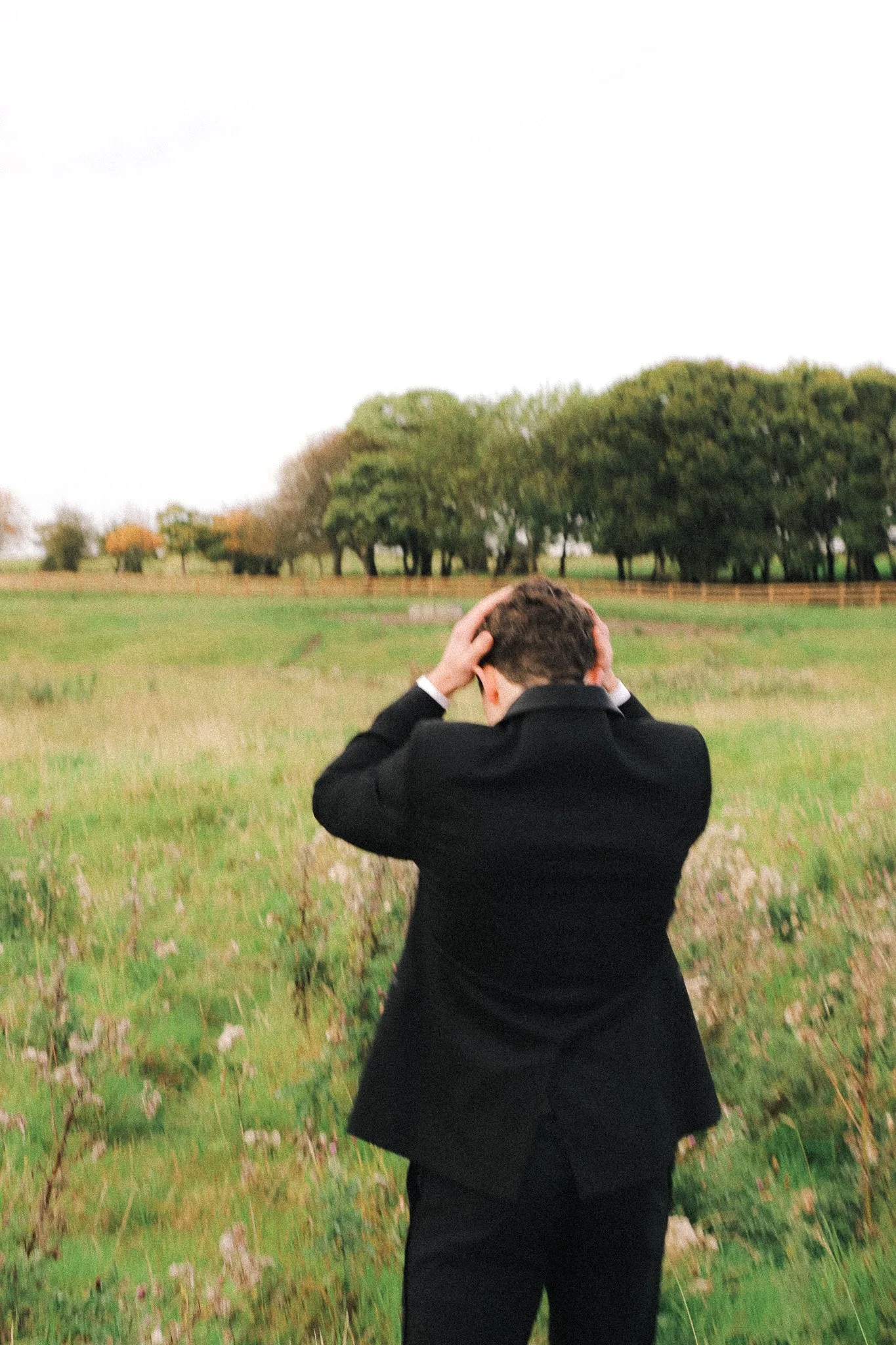 A person dressed in a black suit standing in a grassy field, facing away, with hands on their head and looking down, with trees in the background.