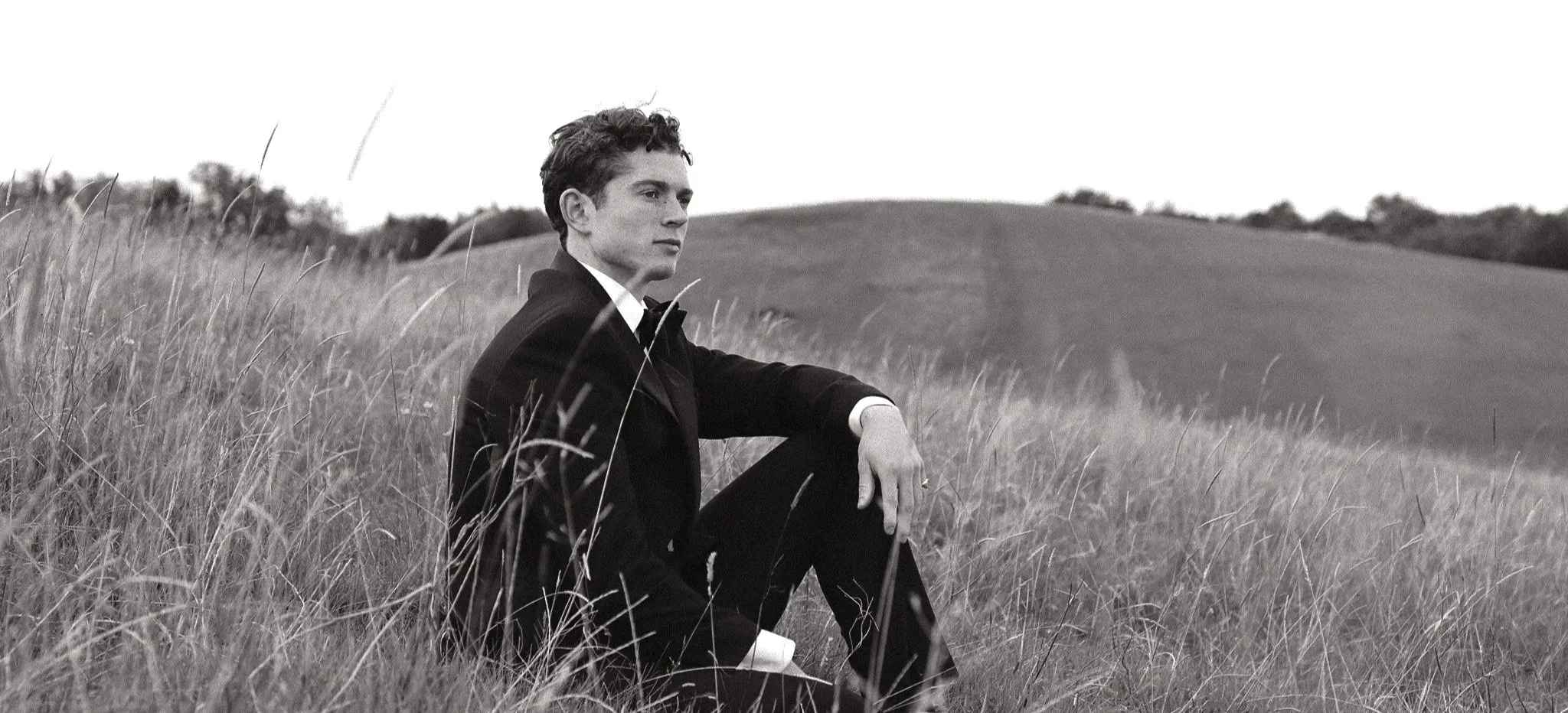 A young man in a suit sitting in a grassy field, looking contemplative, with rolling hills in the background in black and white.
