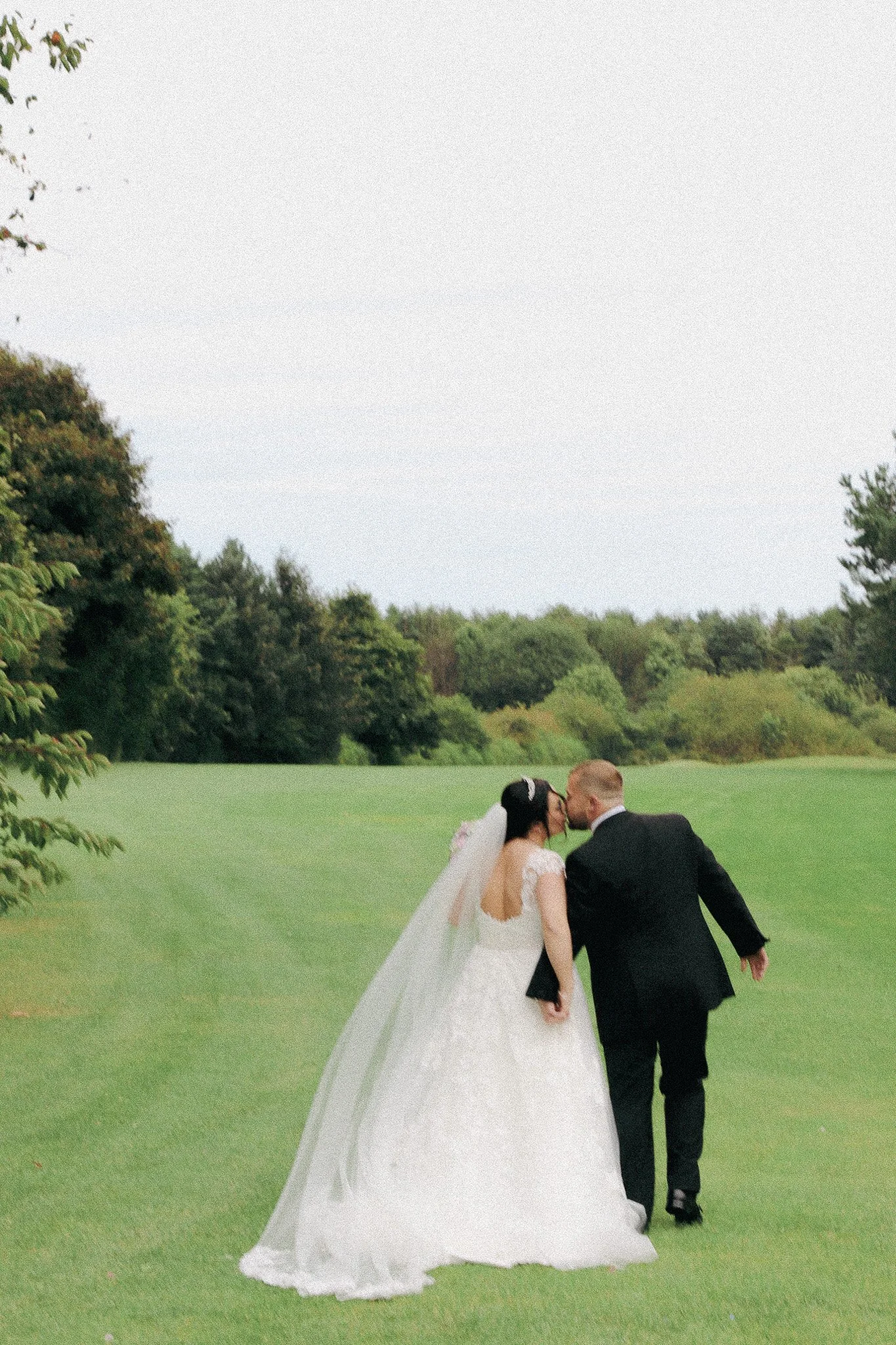 A bride and groom sharing a kiss outside on a grassy field with trees in the background.