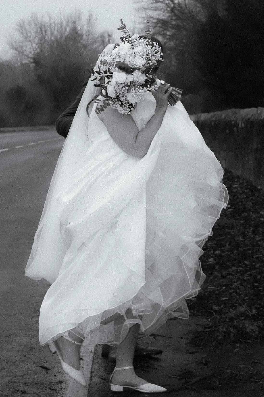 A bride in a white wedding dress holding a bouquet of flowers, standing on a rural road with trees in the background.