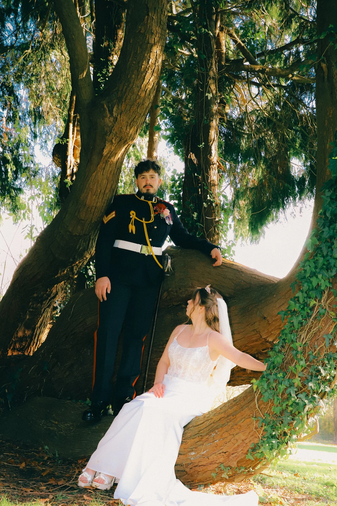 A man in a military uniform standing beside a woman in a wedding dress sitting on a large tree branch, outdoors under a large tree.
