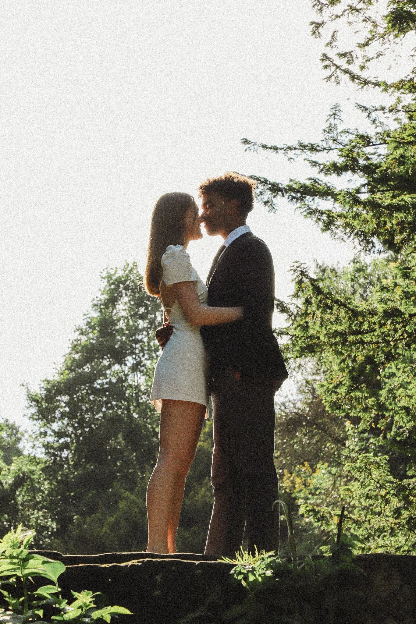 A silhouette of a couple, a woman in a white dress and a man in a black suit, standing close together outdoors with trees around them at sunset.