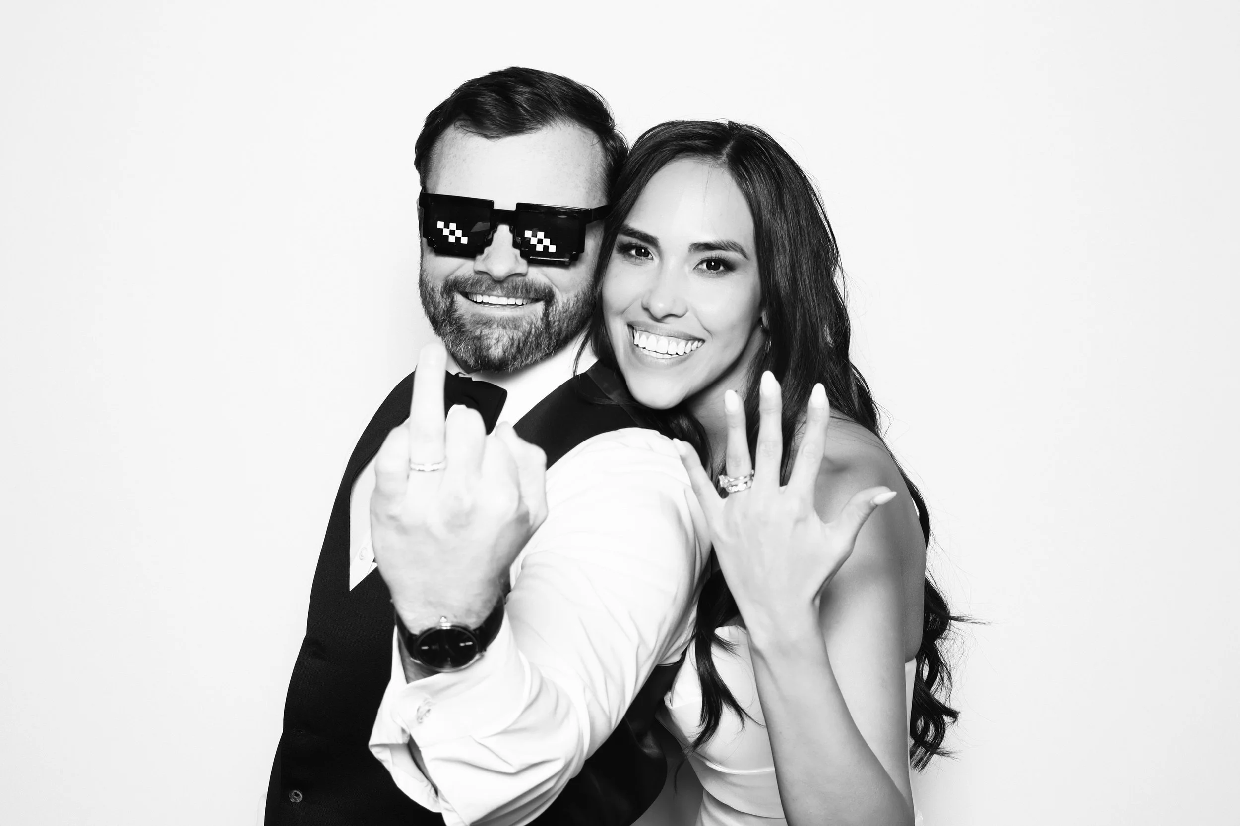 A smiling man in a tuxedo with pixelated sunglasses and a woman in a white dress posing together, showing peace signs on a plain white background.