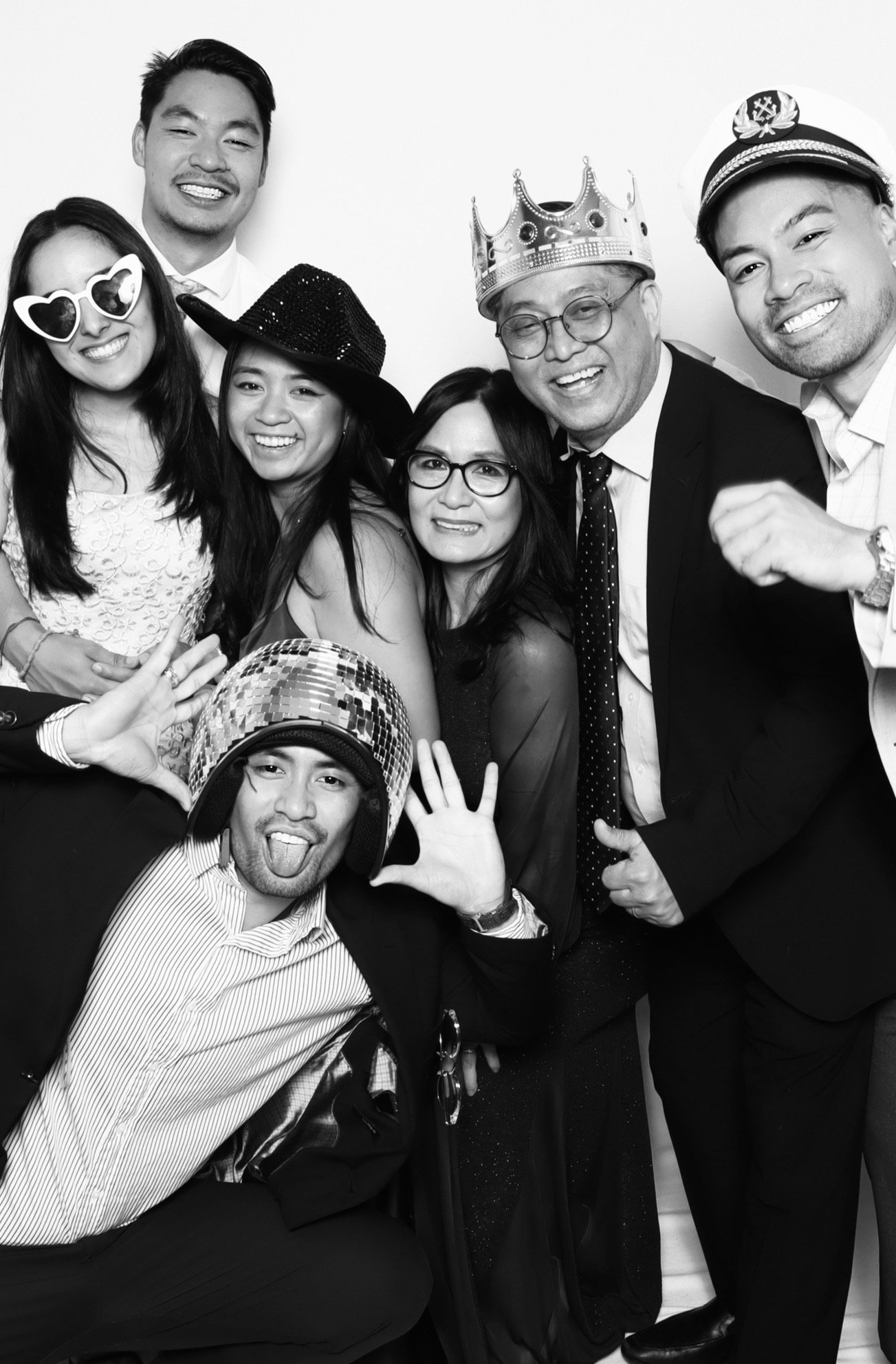 Group of people celebrating, wearing costumes and accessories like hats, sunglasses, and a crown, in a photo booth with a white background.