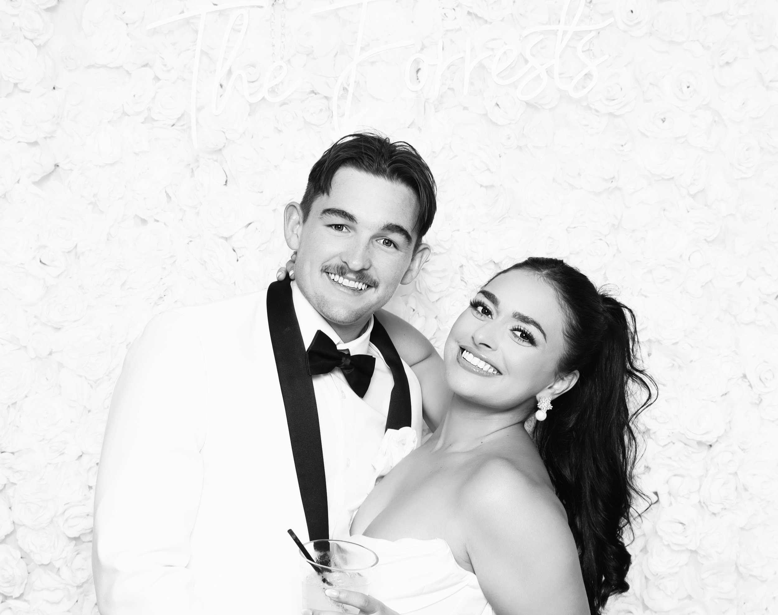 A happy man in a white suit with a bowtie and a woman with long hair in a strapless dress posing together at a celebration, with a floral backdrop and a neon sign.
