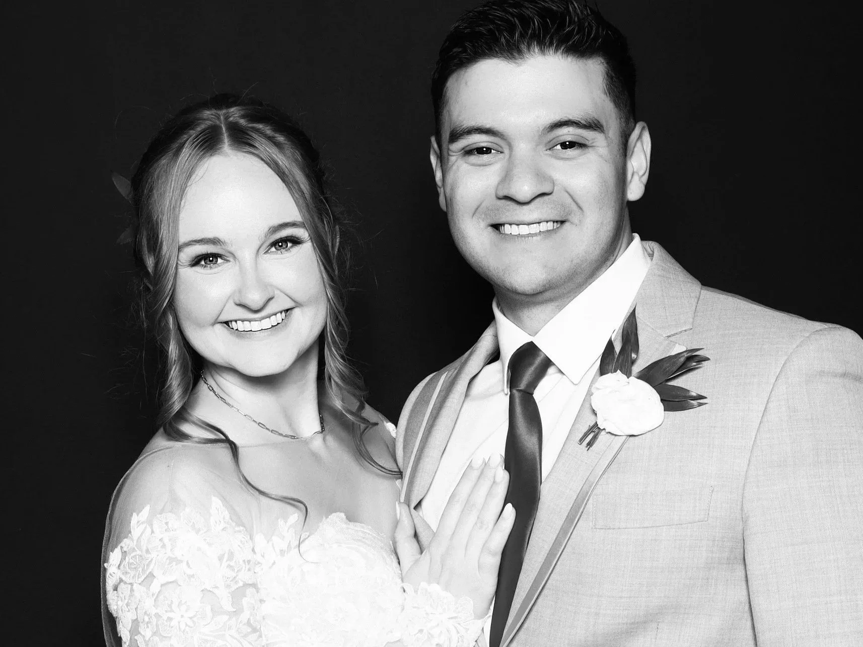 Black and white photo of a smiling bride and groom dressed in wedding attire, standing close together against a dark background.