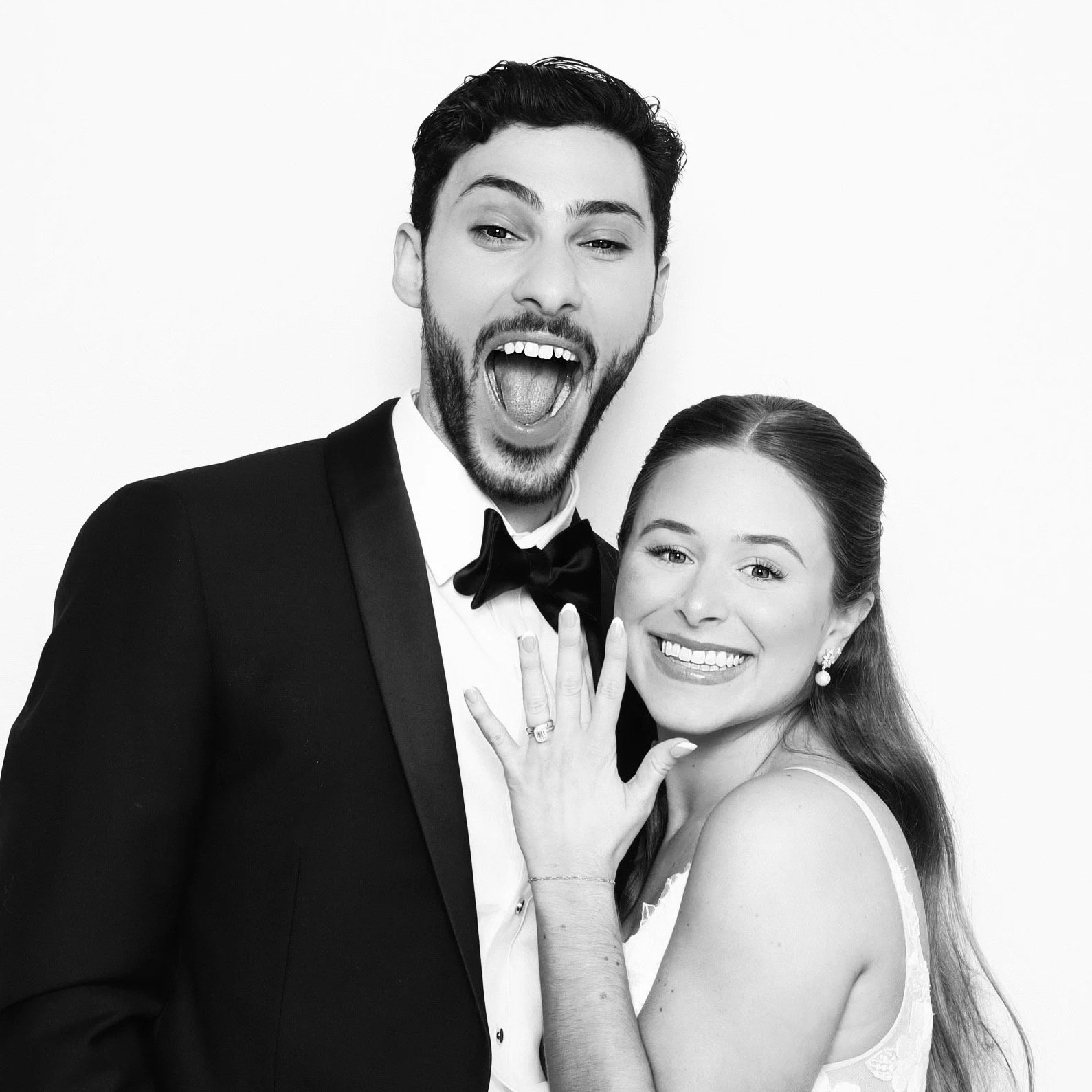 A black-and-white photo of a smiling couple in formal attire, with the man in a tuxedo and the woman in a dress, showing off a ring on her finger.