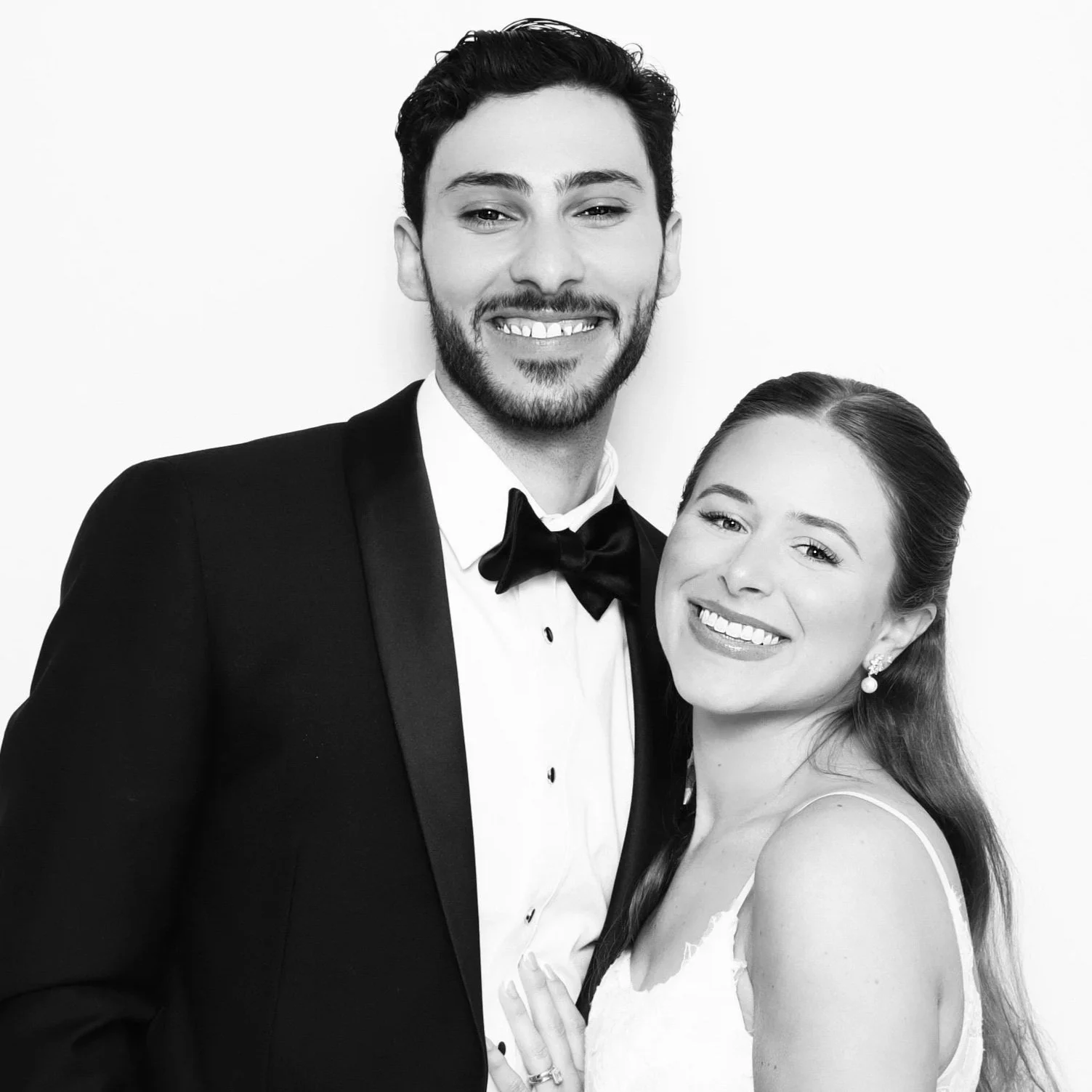 A black and white photo of a smiling couple in formal attire, with the man wearing a tuxedo and the woman wearing a dress and pearl earrings.