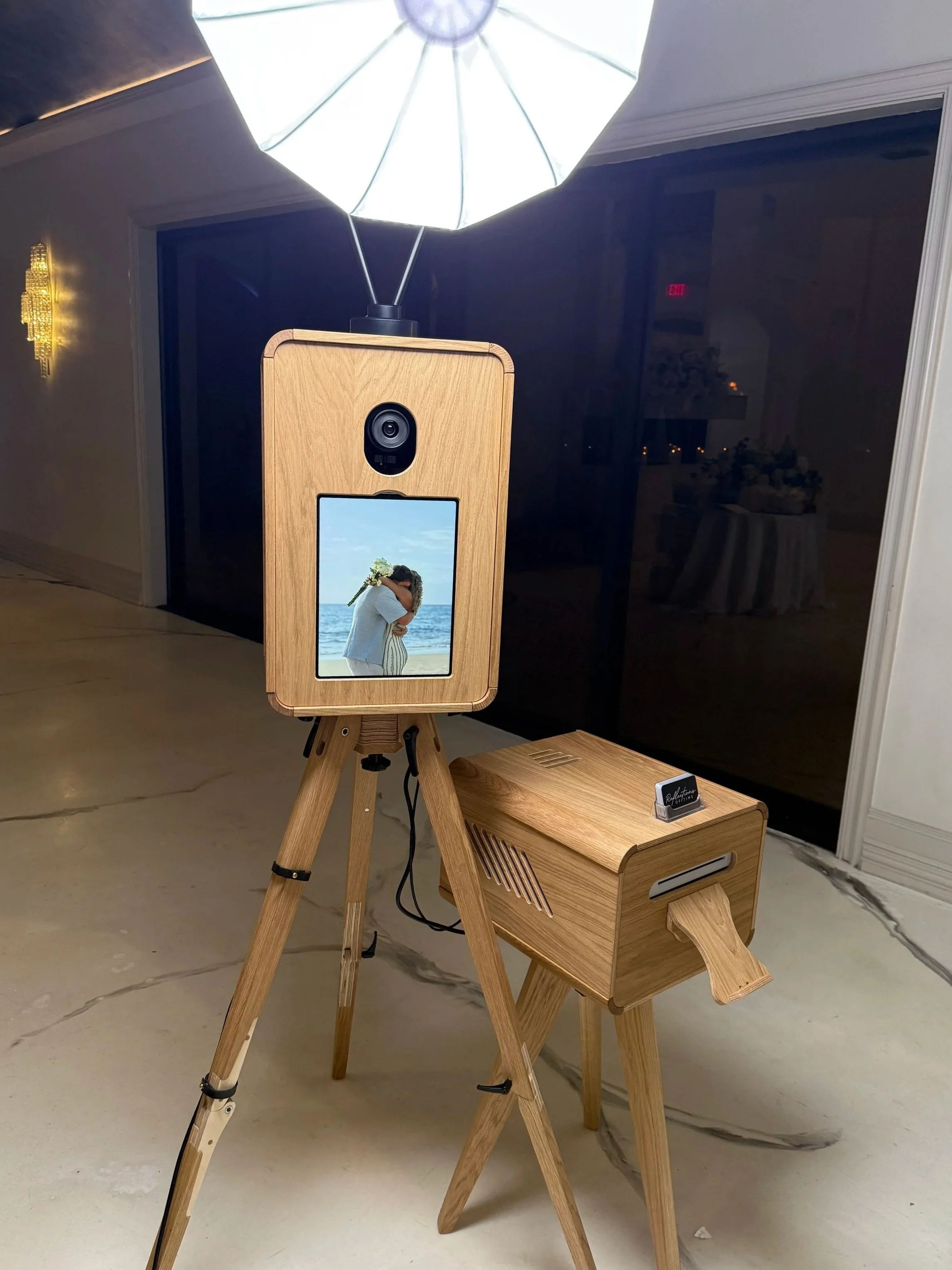 A wooden photo booth with a mirror on the front displaying a couple hugging on a beach, set up indoors next to a black wall and a marble floor.