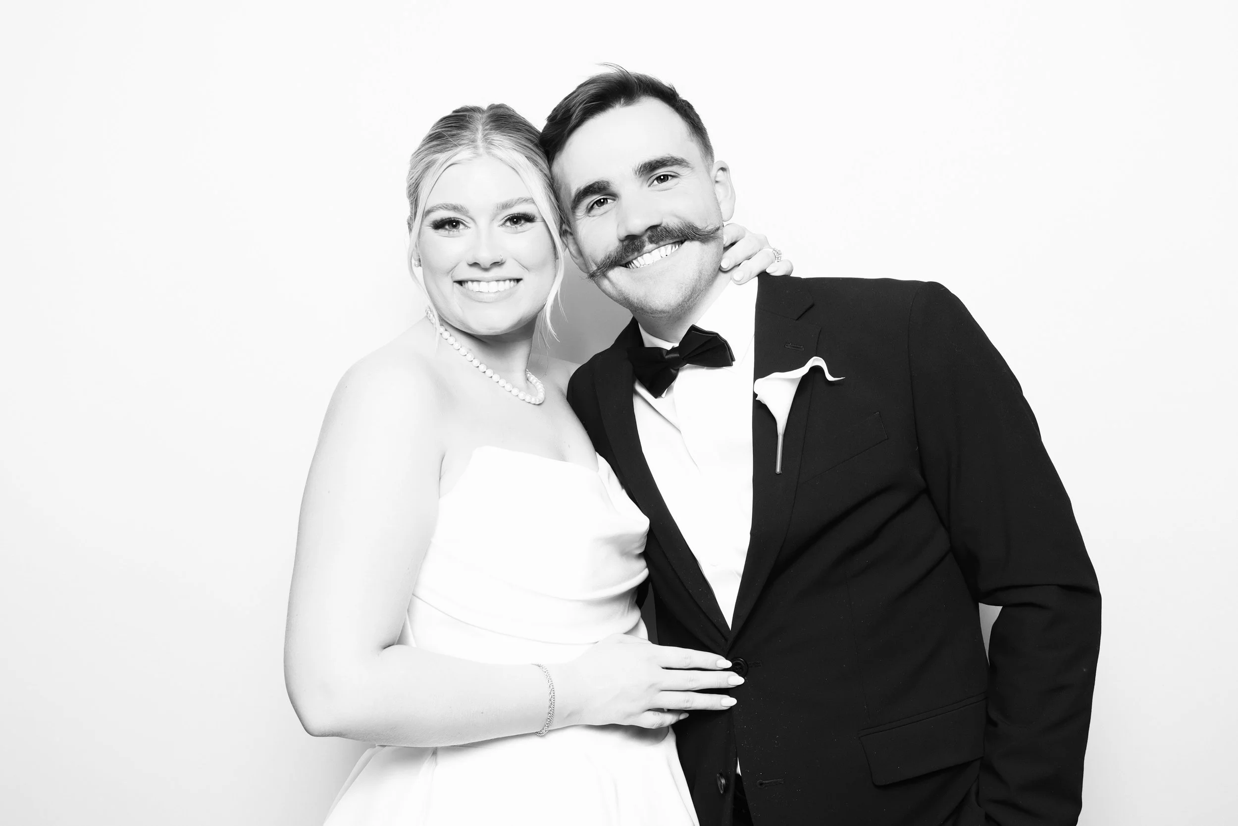 Black and white photo of a smiling bride and groom at their wedding, dressed in formal wedding attire, standing close together against a plain background.