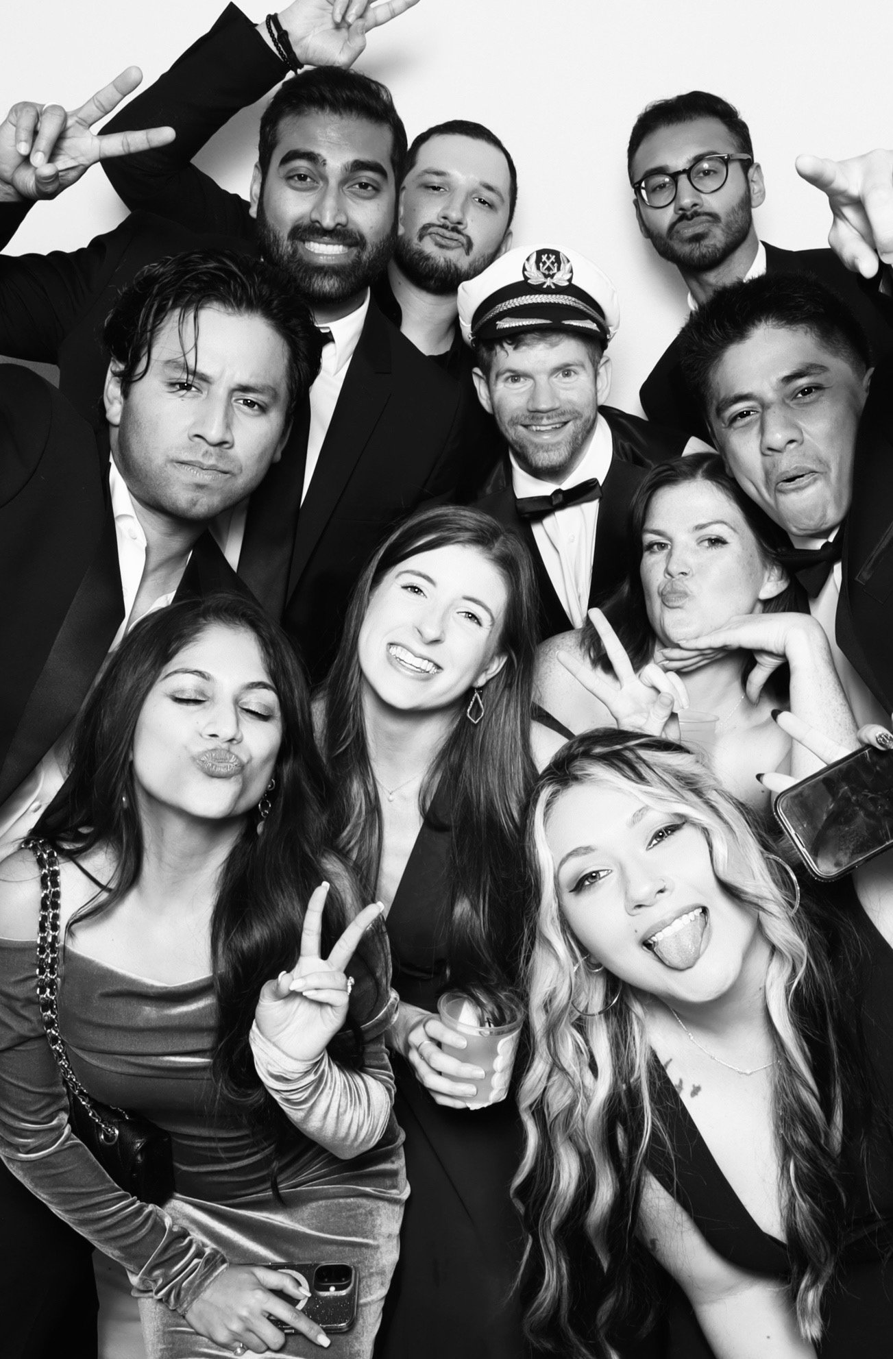 Group of friends taking a lively, black-and-white photo at a party, including women making peace signs and men in suits, some wearing glasses, posing closely together.