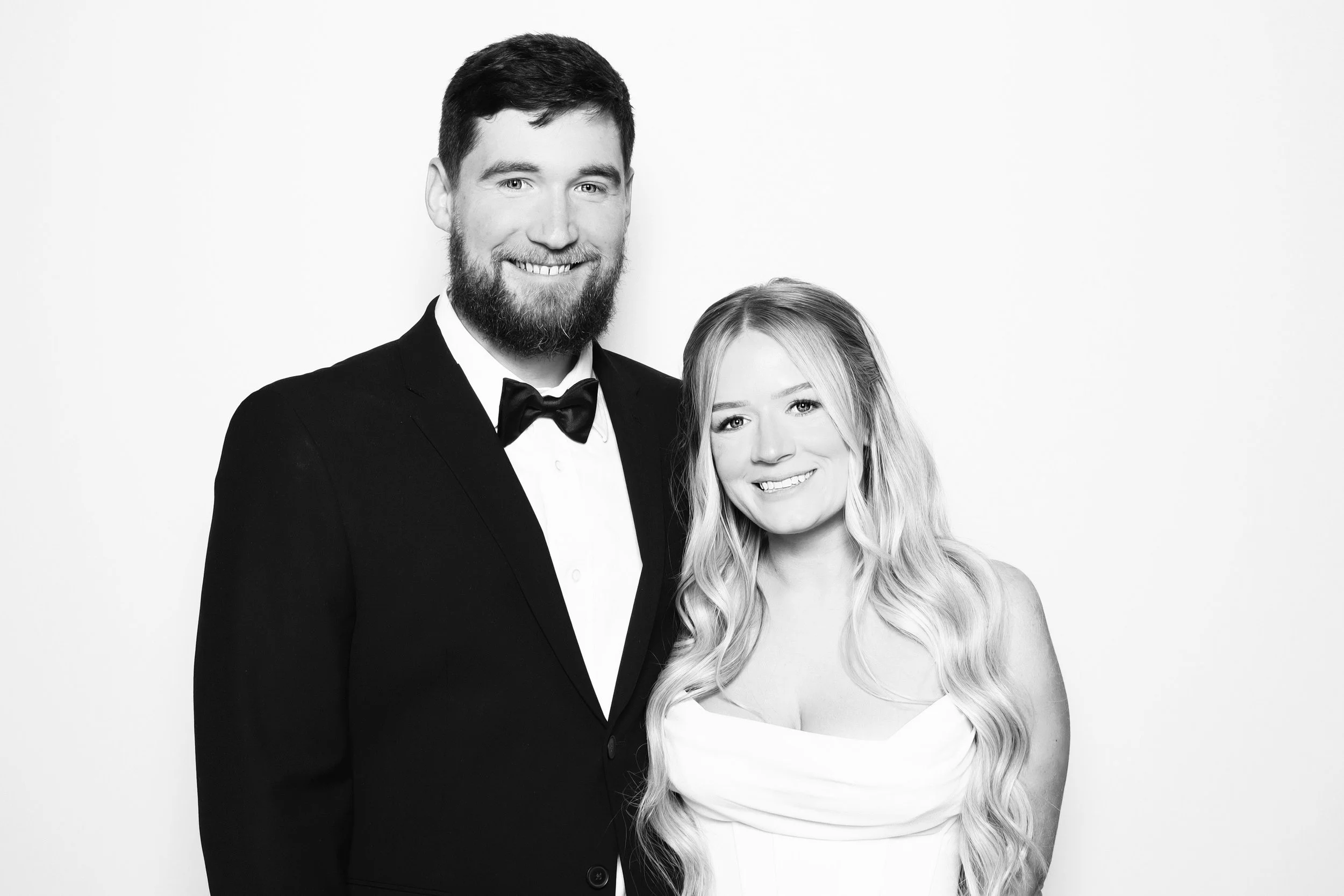 A black-and-white photo of a man in a tuxedo with a bow tie and a woman in a strapless dress, both smiling and standing close together against a plain background.