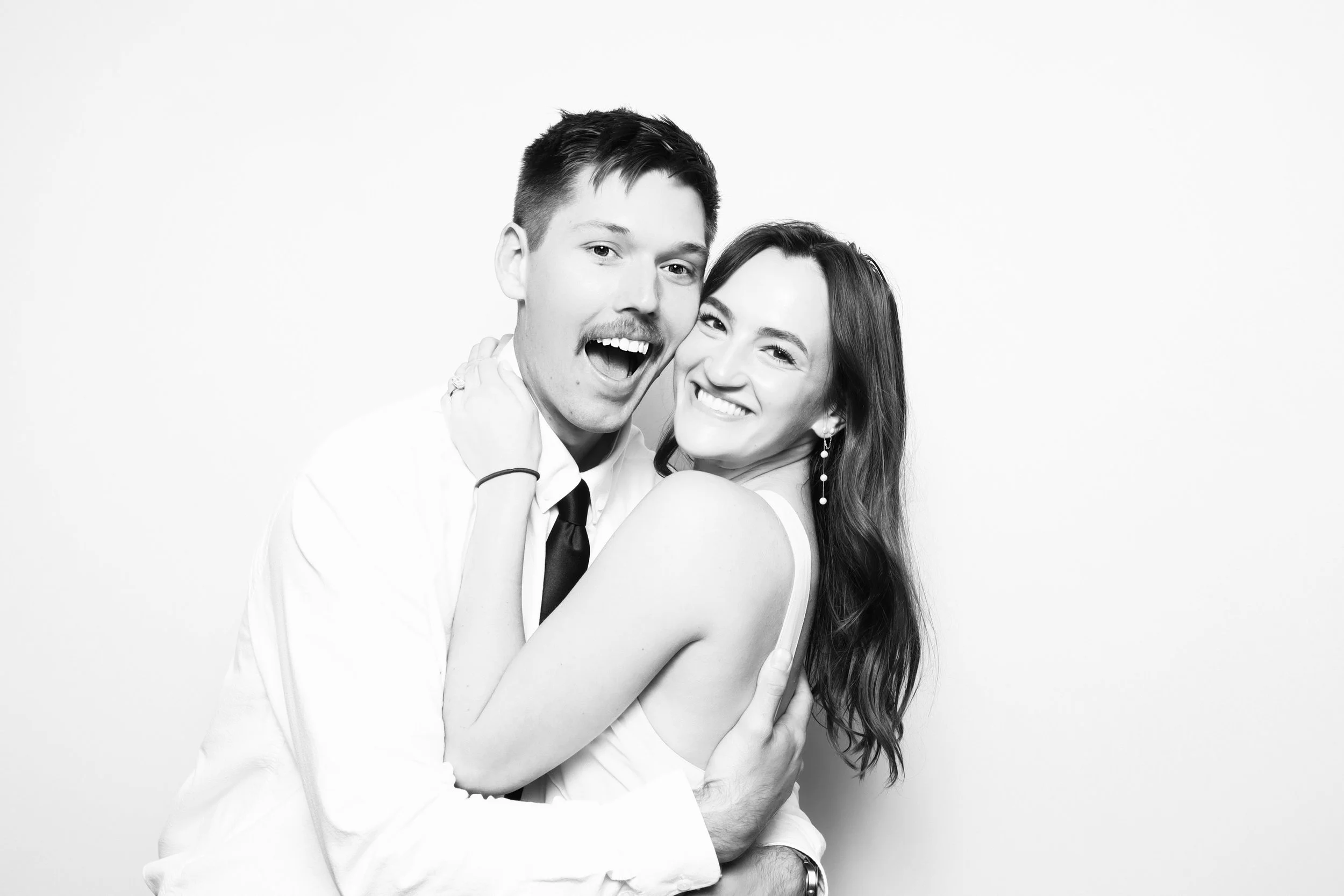 A black and white photo of a smiling young couple embracing each other against a plain background. The man has short dark hair, a mustache, and is wearing a shirt, tie, and watch. The woman has long dark hair, earrings, and is wearing a sleeveless dr