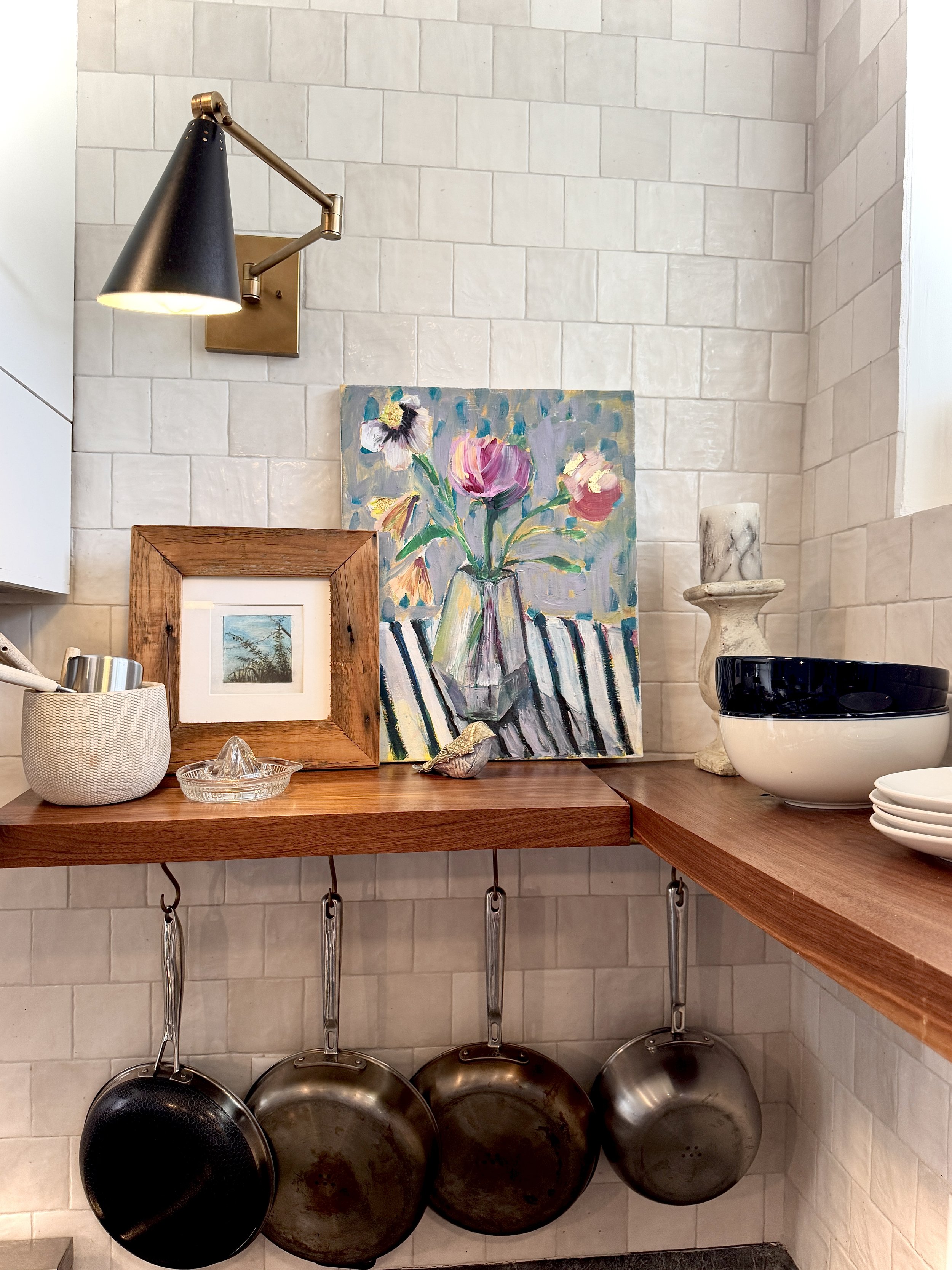 A kitchen wall with beige subway tiles and a wooden countertop. Three large cast iron skillets are hanging beneath the countertop. A black frying pan is hanging on the left side. On the countertop, there is a white textured container with utensils, a