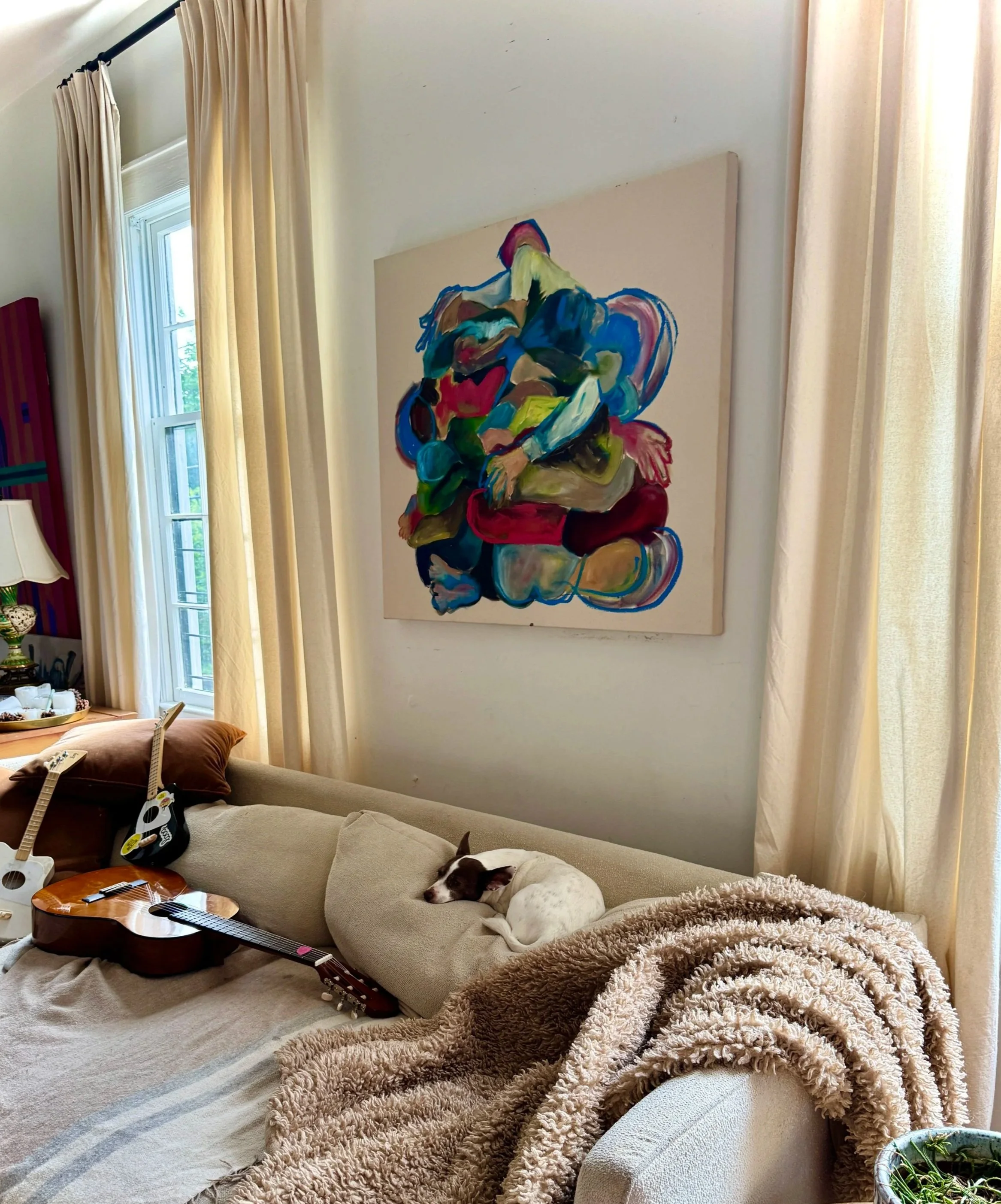 Living room with beige sofa, a sleeping dog, a guitar, a blanket, and a colorful abstract painting on the wall near a window with cream curtains.
