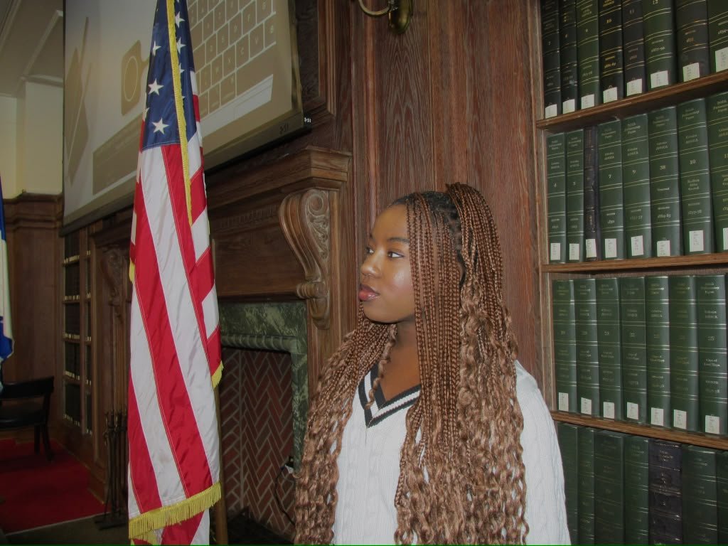 Angel in a wood panelled room surrounded by books and next to the American flag
