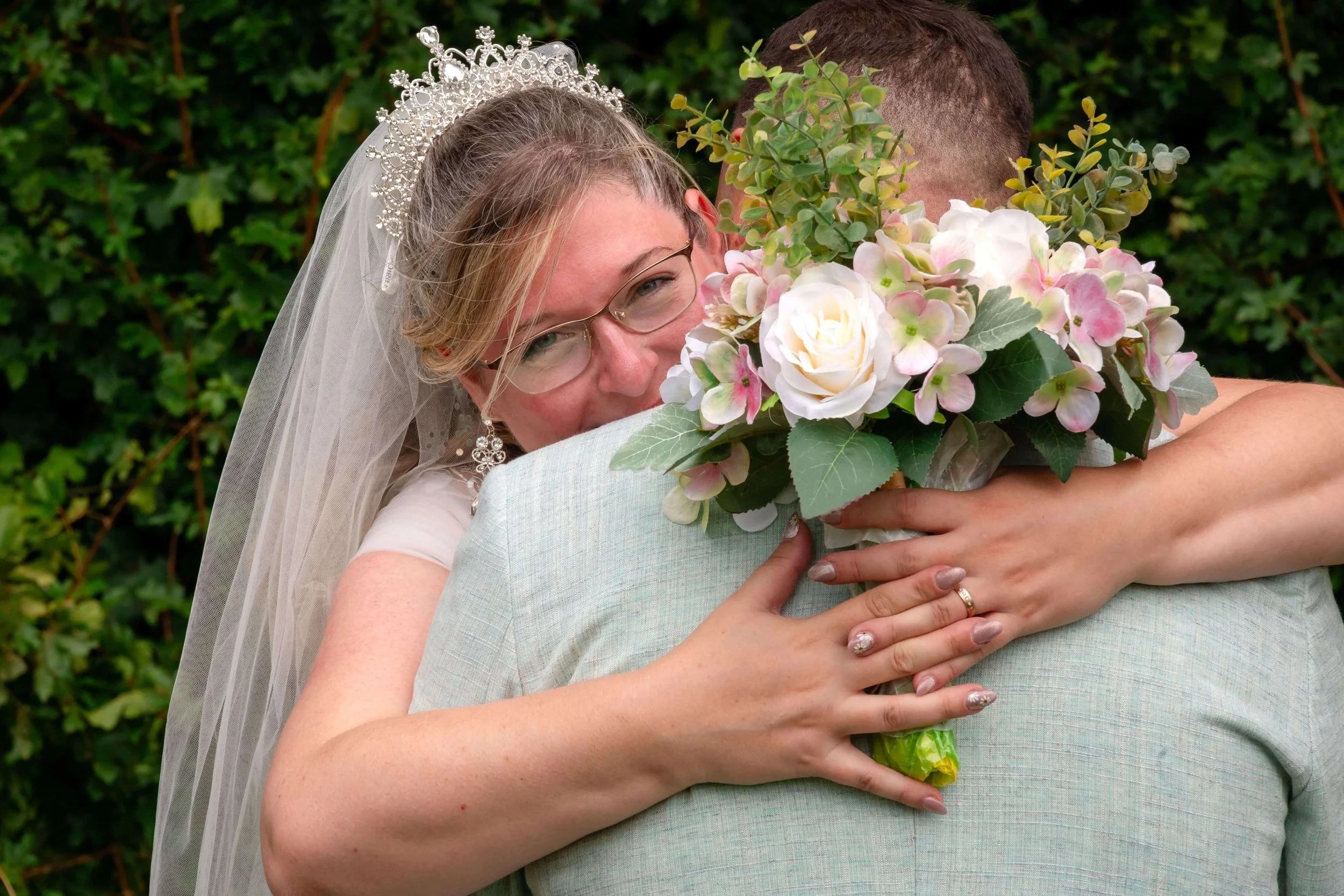 Hochzeitspaar umarmt sich in freier Trauung in Giessen. Die Braut trägt eine Tiara und eine Brille, hält einen Blumenstrauß, der nur teilweise sichtbar ist, während der Bräutigam mit seinem Rücken zur Kamera steht. Im Hintergrund grüne Büsche.