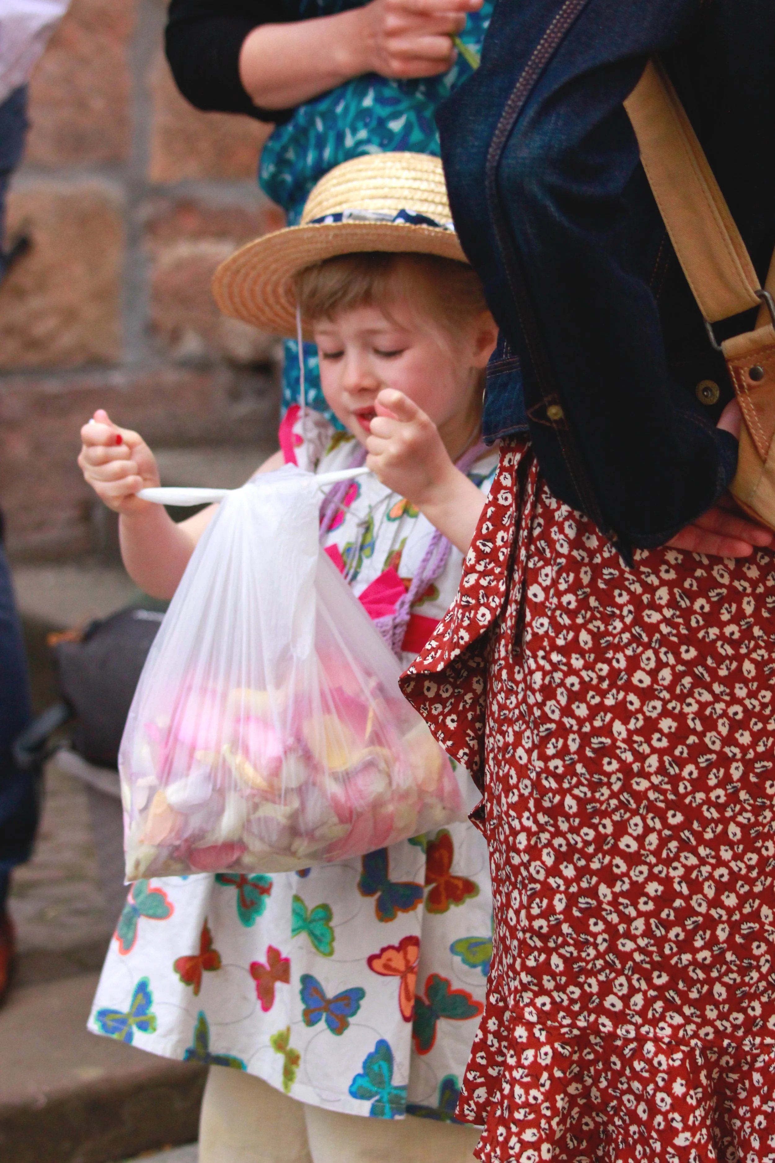 Ein junges Mädchen trägt einen Strohhut und ein Kleid mit Schmetterlingen, während sie in einer Plastiktüte Süßigkeiten hält. Es sind andere Menschen um sie herum, eine Person trägt eine rote Hose mit weißen Punkten. in ihrer Hochzeit in Marburg