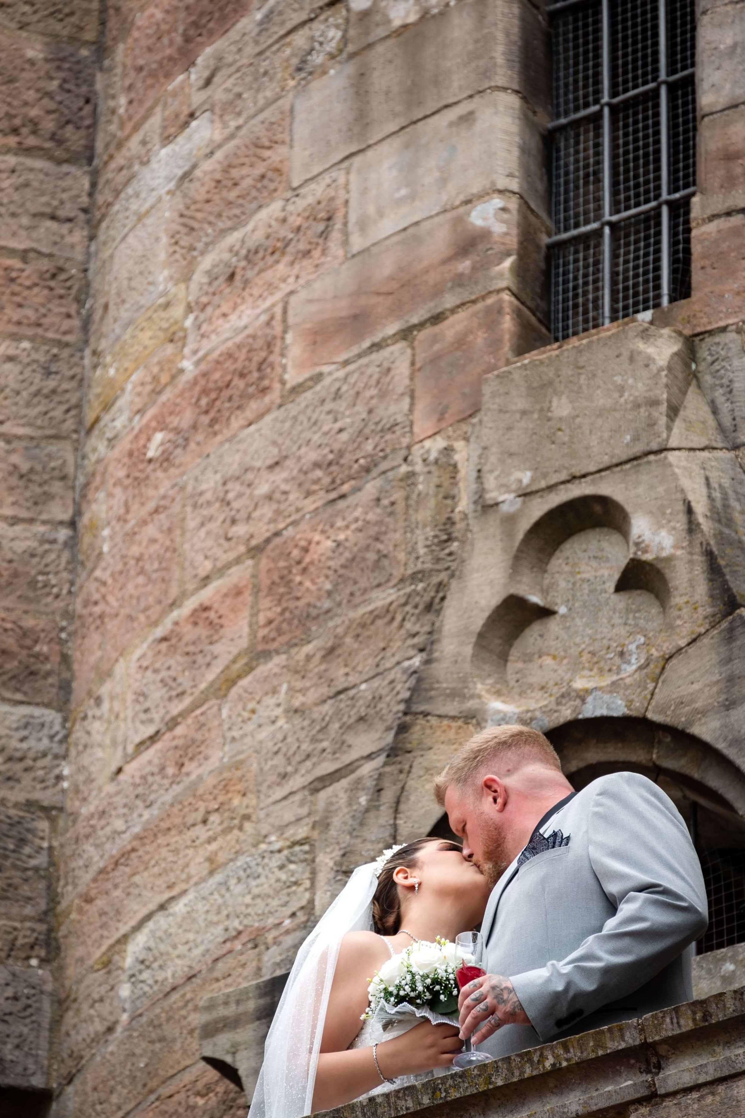 Ein Brautpaar küsst sich auf einem Balkon vor einer alten Steinmauer mit spitzbogigen Fenstern, Hochzeit in Marburg