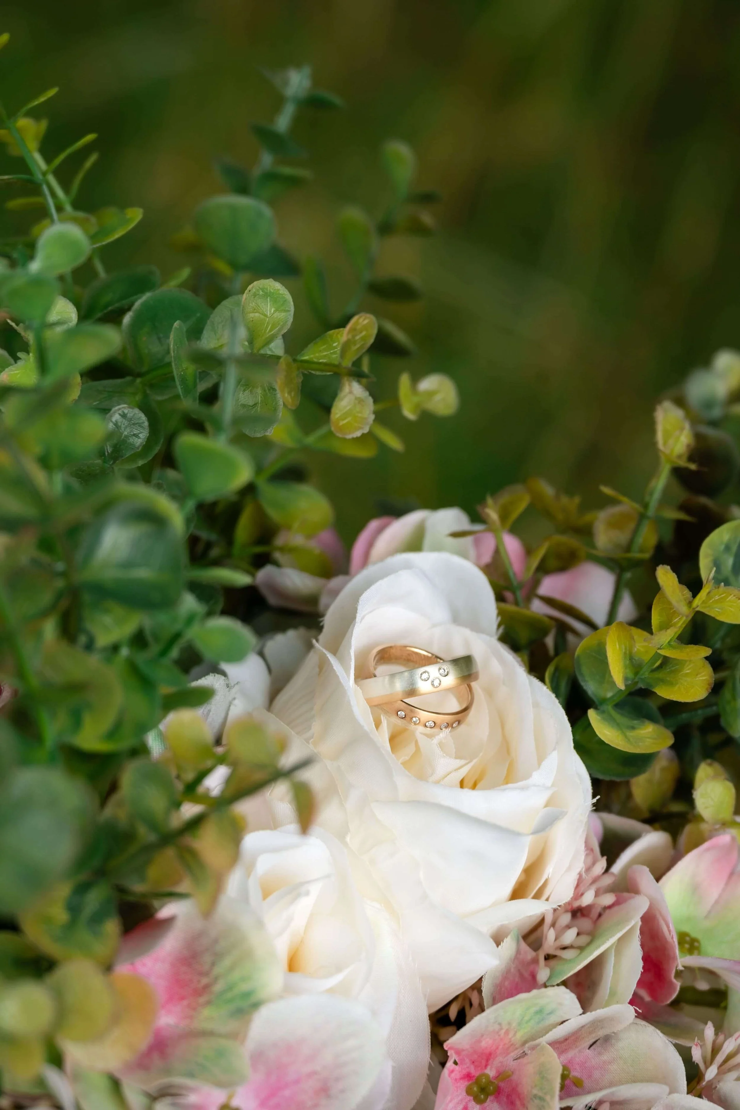 Eheringe liegen in einer weißen Rose, umgeben von grünen Blättern und pink-weißen Blüten.