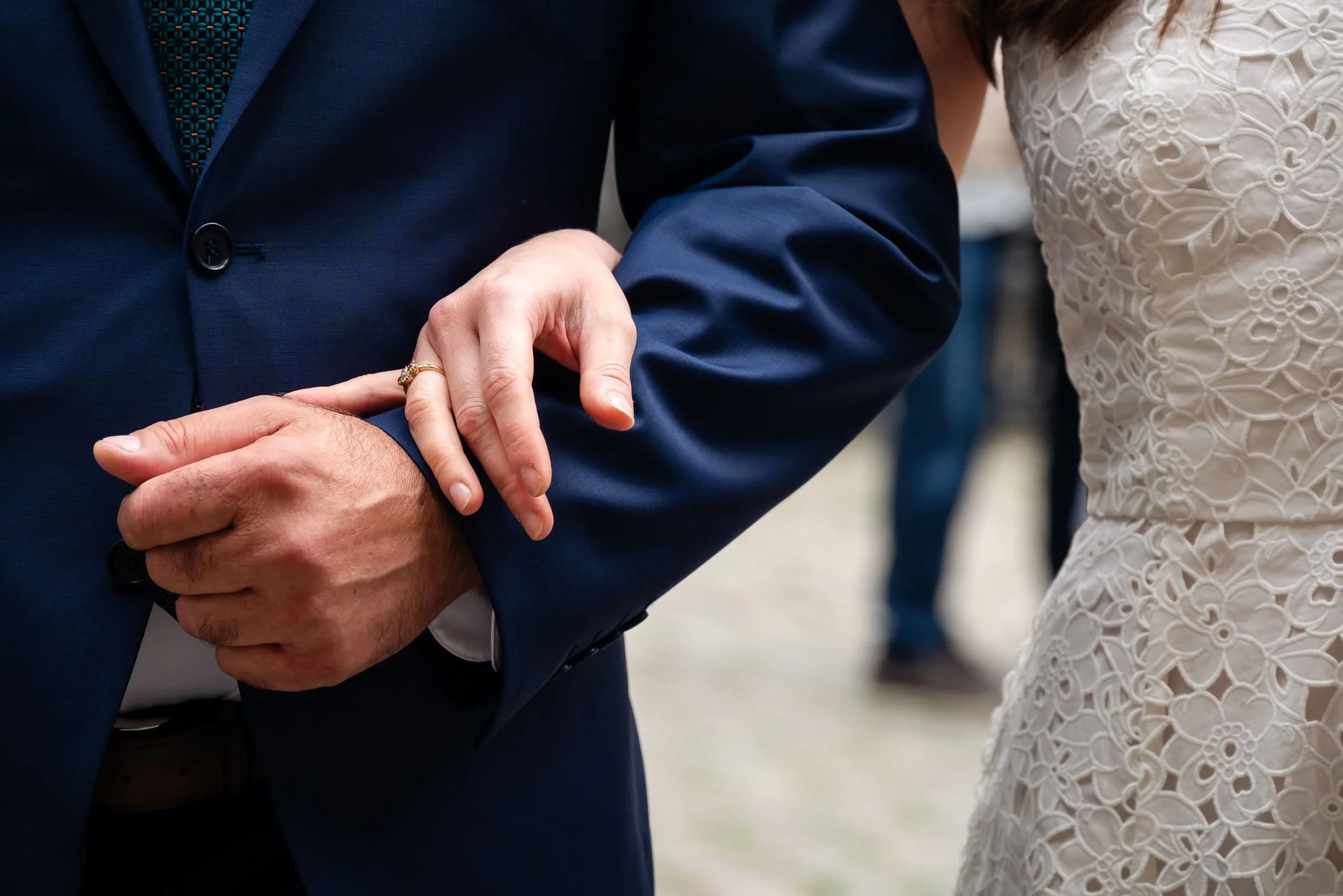 Nahaufnahme eines Paares bei einer Hochzeit, die Frau hält den Arm des Mannes an seinem Ärmel, beide tragen formelle Kleidung, die Frau in einem weißen Kleid mit Stickerei, der Mann in einem blauen Anzug.