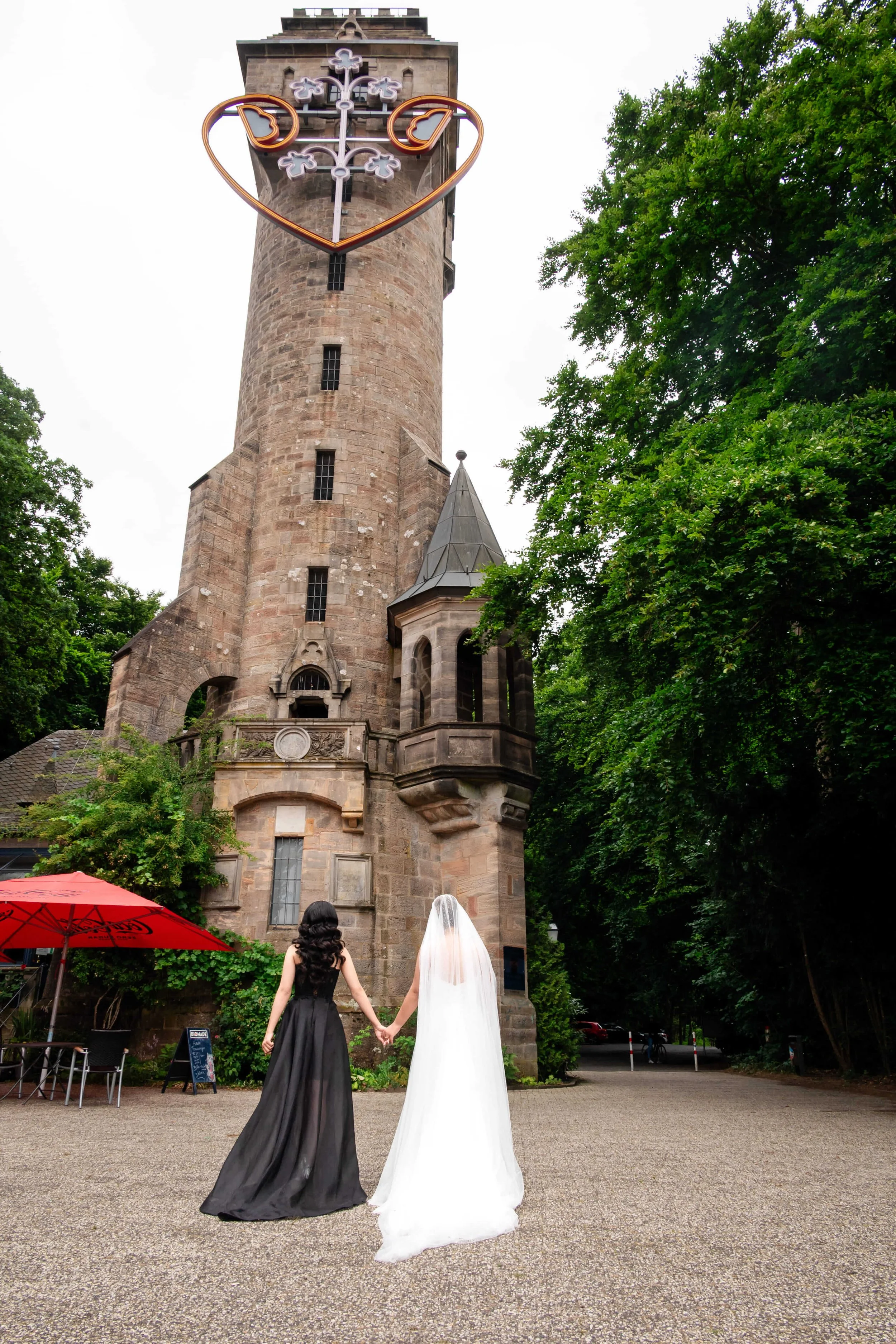 Zwei Frauen, eine in einem schwarzen Kleid, die andere in einem weißen Brautkleid, halten sich an den Händen vor einem alten Turm mit einem Neon-Herz und Kreuz oben. Das Bild ist im Freien, mit Bäumen auf der rechten Seite in Marburg