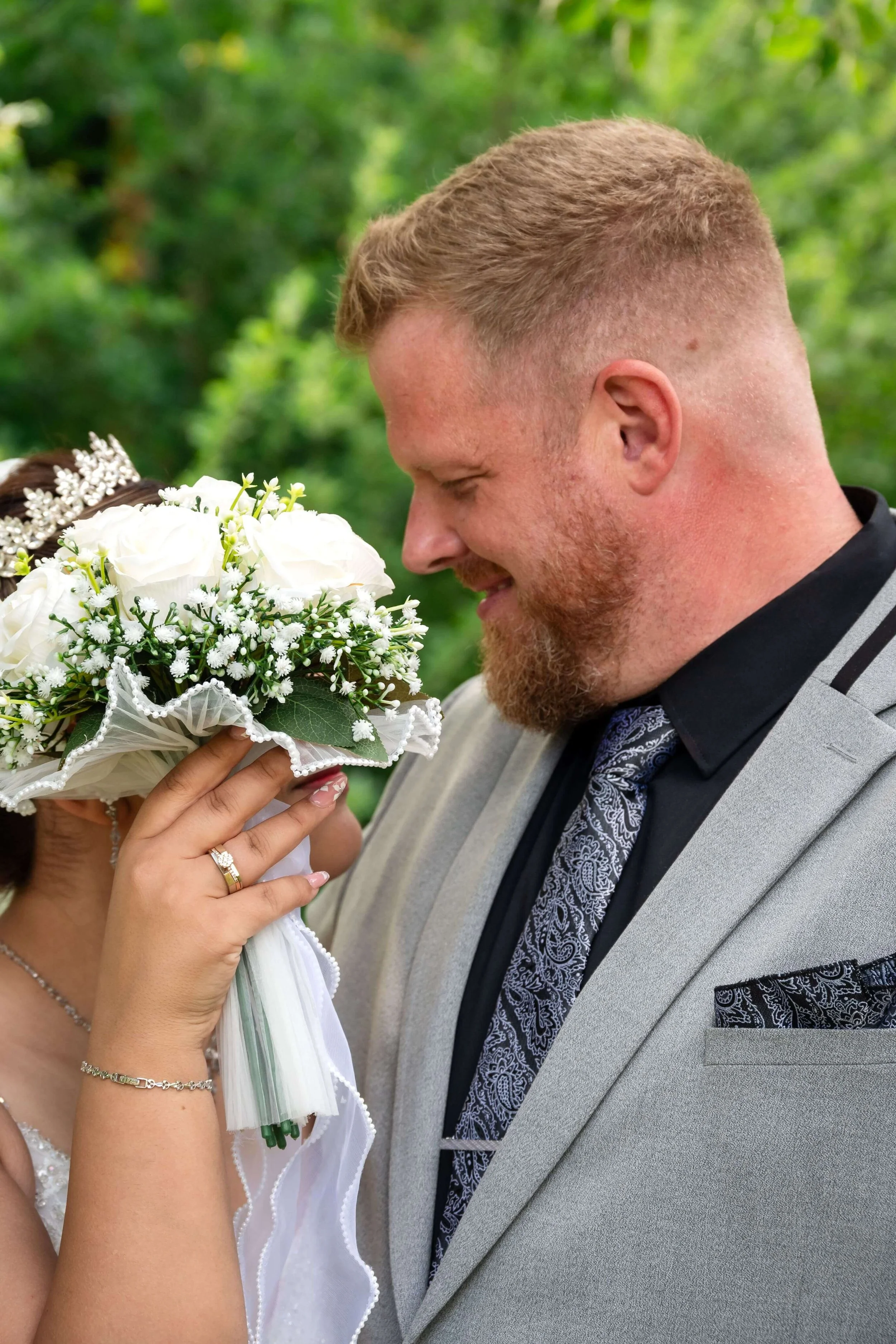 Ein Brautpaar bei einer Hochzeit in Marburg, der Bräutigam trägt einen grauen Anzug, die Braut hält einen Blumenstrauß vor ihr Gesicht, umarmt den Bräutigam, im Hintergrund grüner Wald 