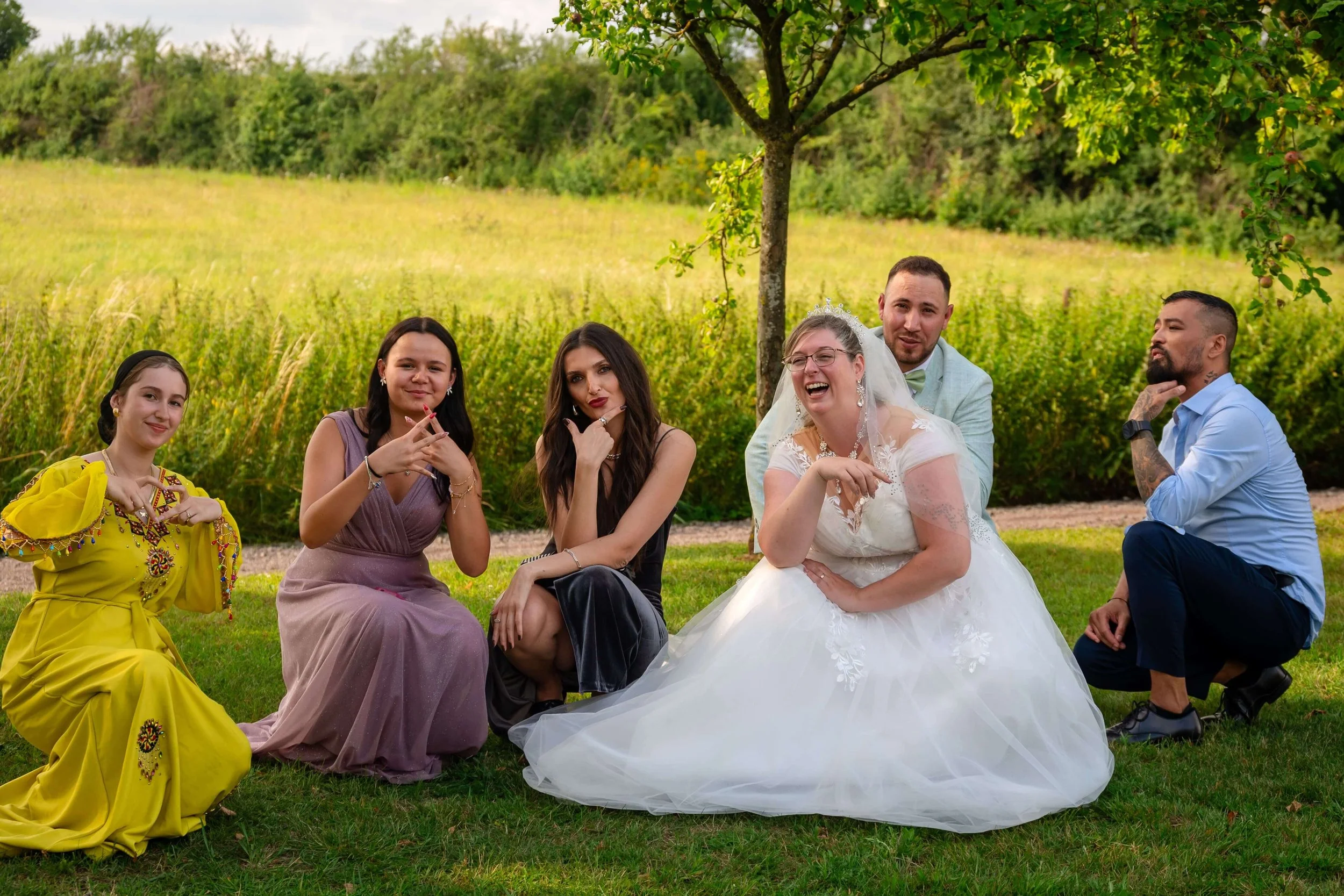 Eine Gruppe von sieben Menschen, darunter eine Braut in weißem Kleid und ein Bräutigam in einem Anzug, sitzen im Freien unter einem Baum, lachen und posieren für ein Gruppenfoto in freier Trauung in Giessen