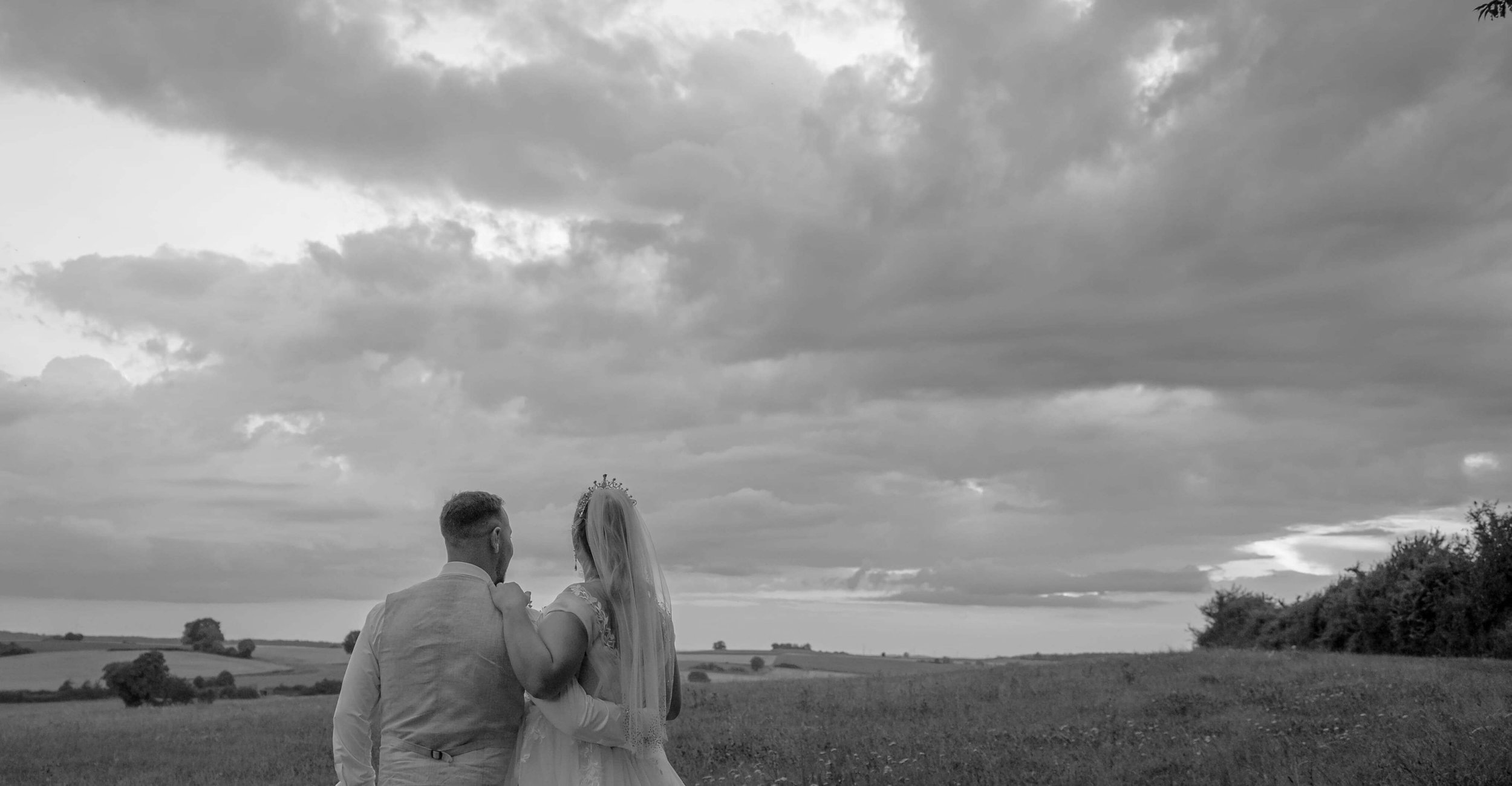Ein Brautpaar steht in einer offenen Landschaft bei bewölktem Himmel, die Frau trägt ein Brautkleid mit Schleier, die Szene ist von einer Hochzeit in Biedenkopf in Schwarz-Weiß.