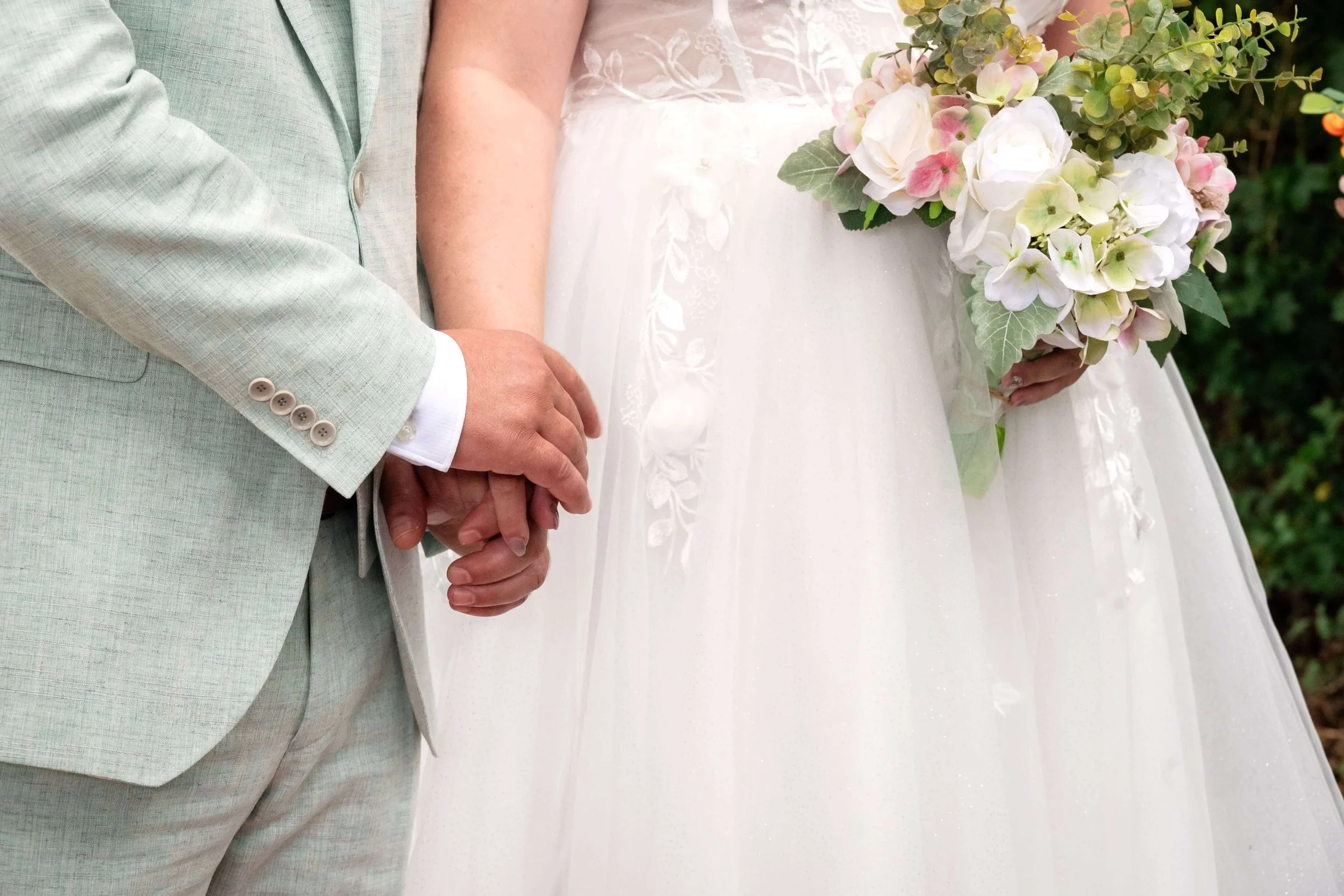 Ein Brautpaar hält sich bei einer Hochzeit an den Händen, die Braut trägt ein weißes Kleid und hält einen Blumenstrauß, der Bräutigam trägt einen hellgrünen Anzug. om ihrer Hochzeit in Marburg