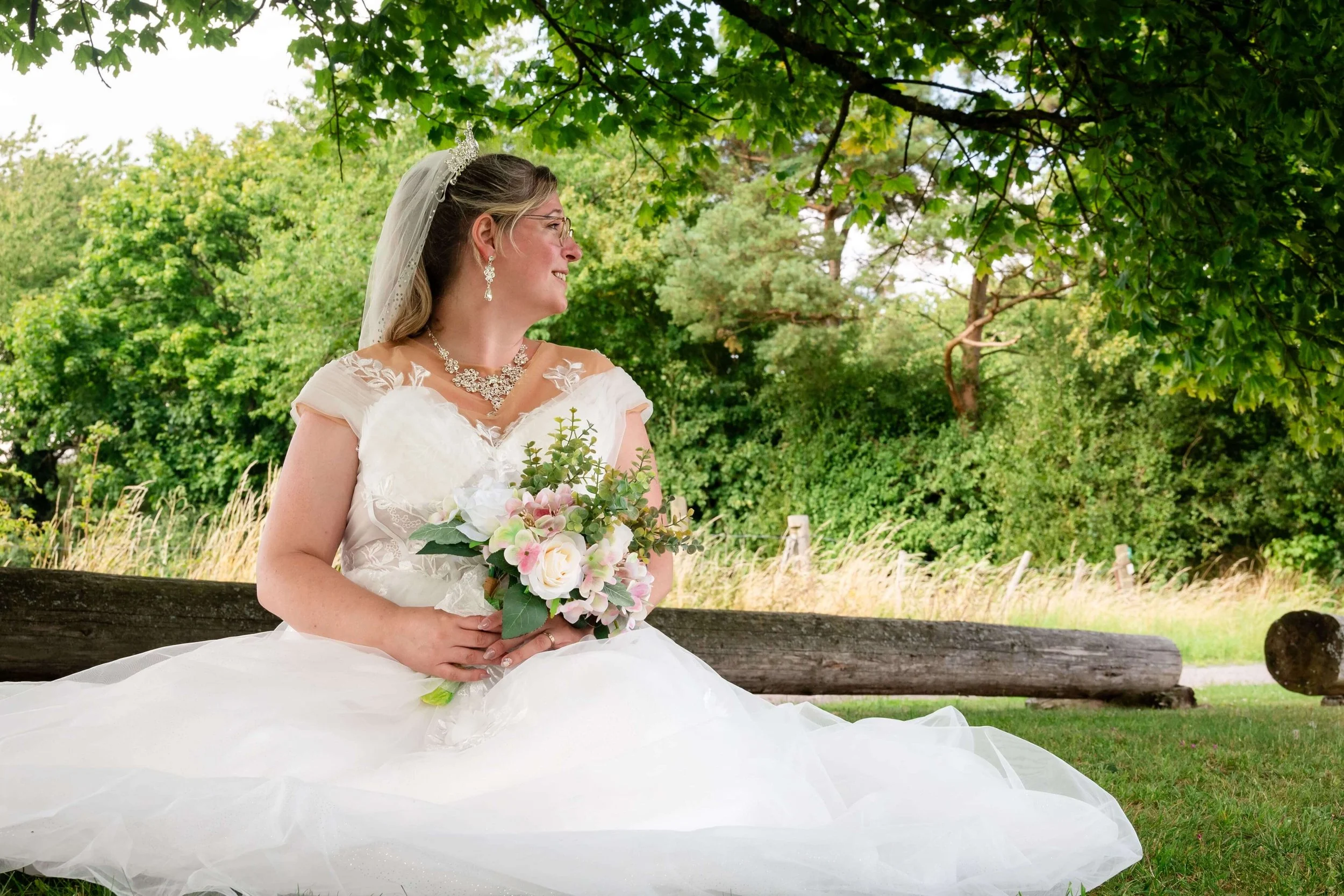 Braut in Hochzeitskleid mit Blume in der Hand sitzt im Grünen under Baum auf Wiese in Marburg.