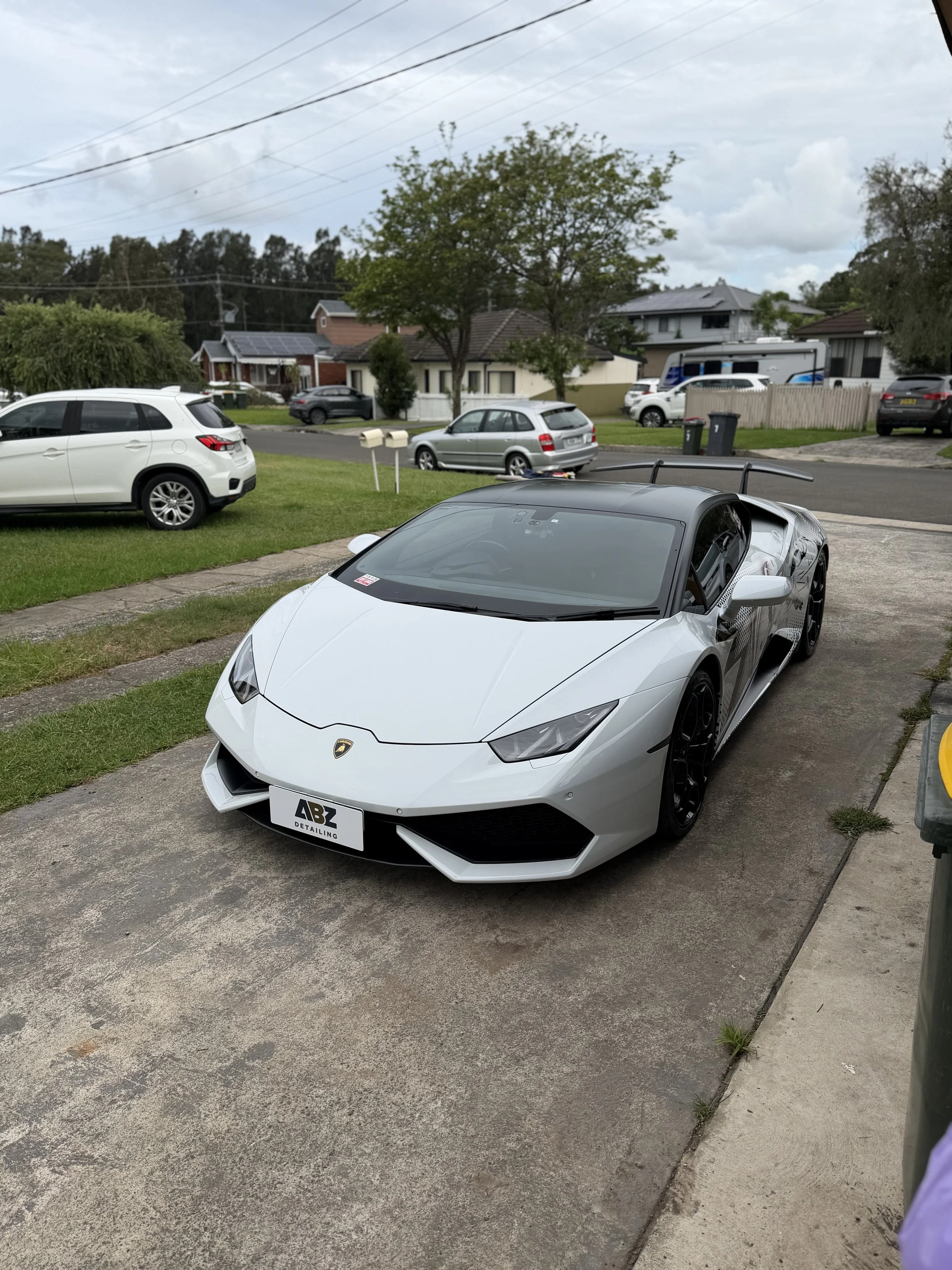 A white Lamborghini sports car parked on a concrete driveway in front of a yellow house with a brown roof, under a blue sky with scattered clouds, casting a shadow on the street.