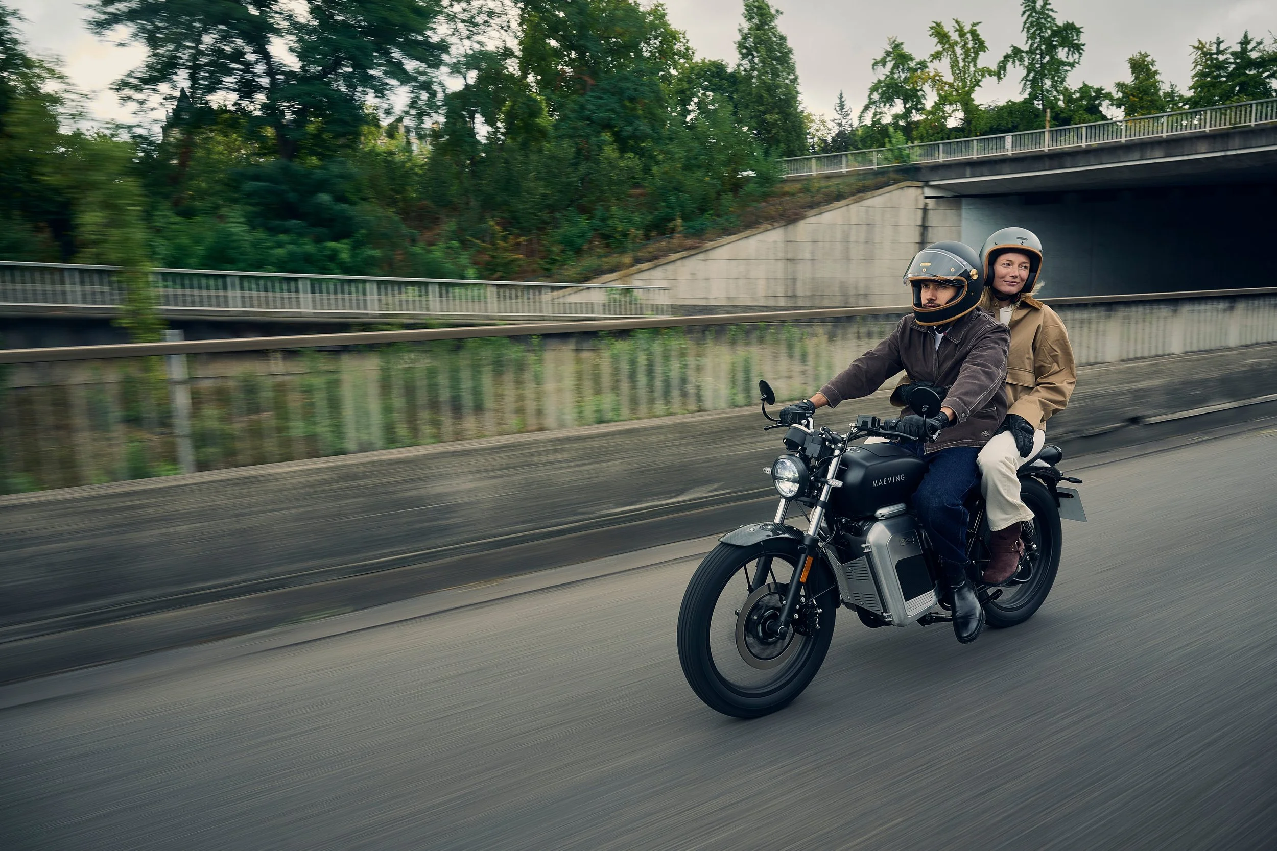 Deux personnes portant des casques de moto, dont un homme et une femme, roulant en moto électrique sur une voie rapide avec de la verdure en arrière-plan.