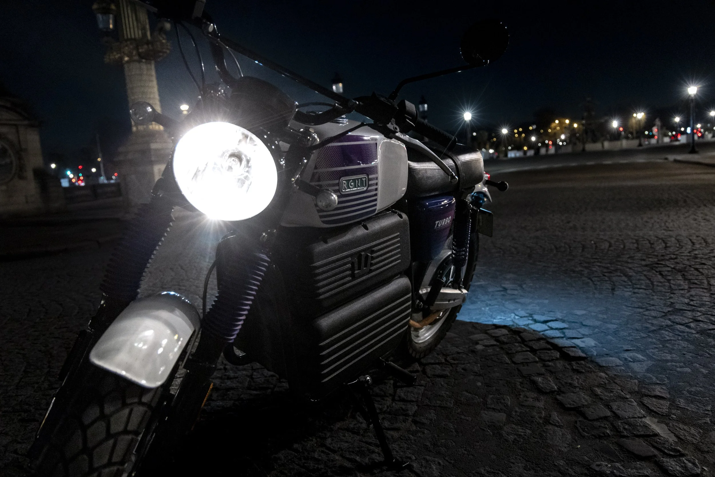 Moto électrique garée sur une place pavée de nuit, avec la lumière du phare allumée et un paysage urbain en arrière-plan.
