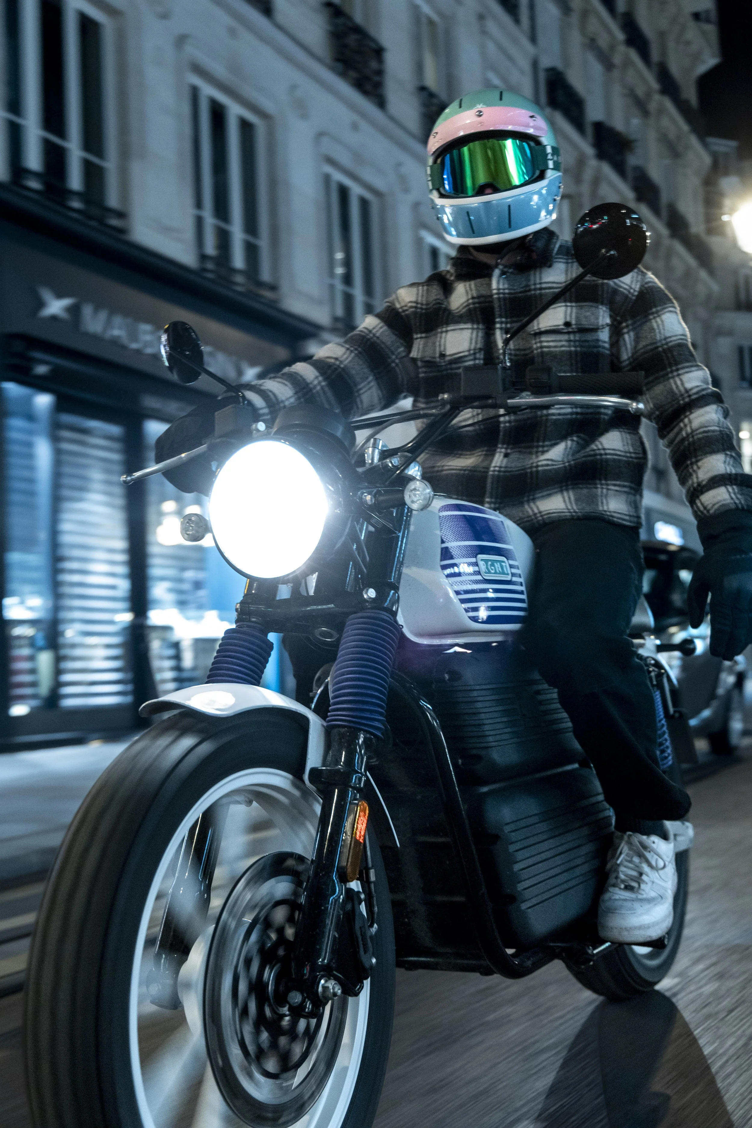 Personne portant un casque de moto électrique coloré et une veste à carreaux, roulant à moto électrique la nuit dans une ville