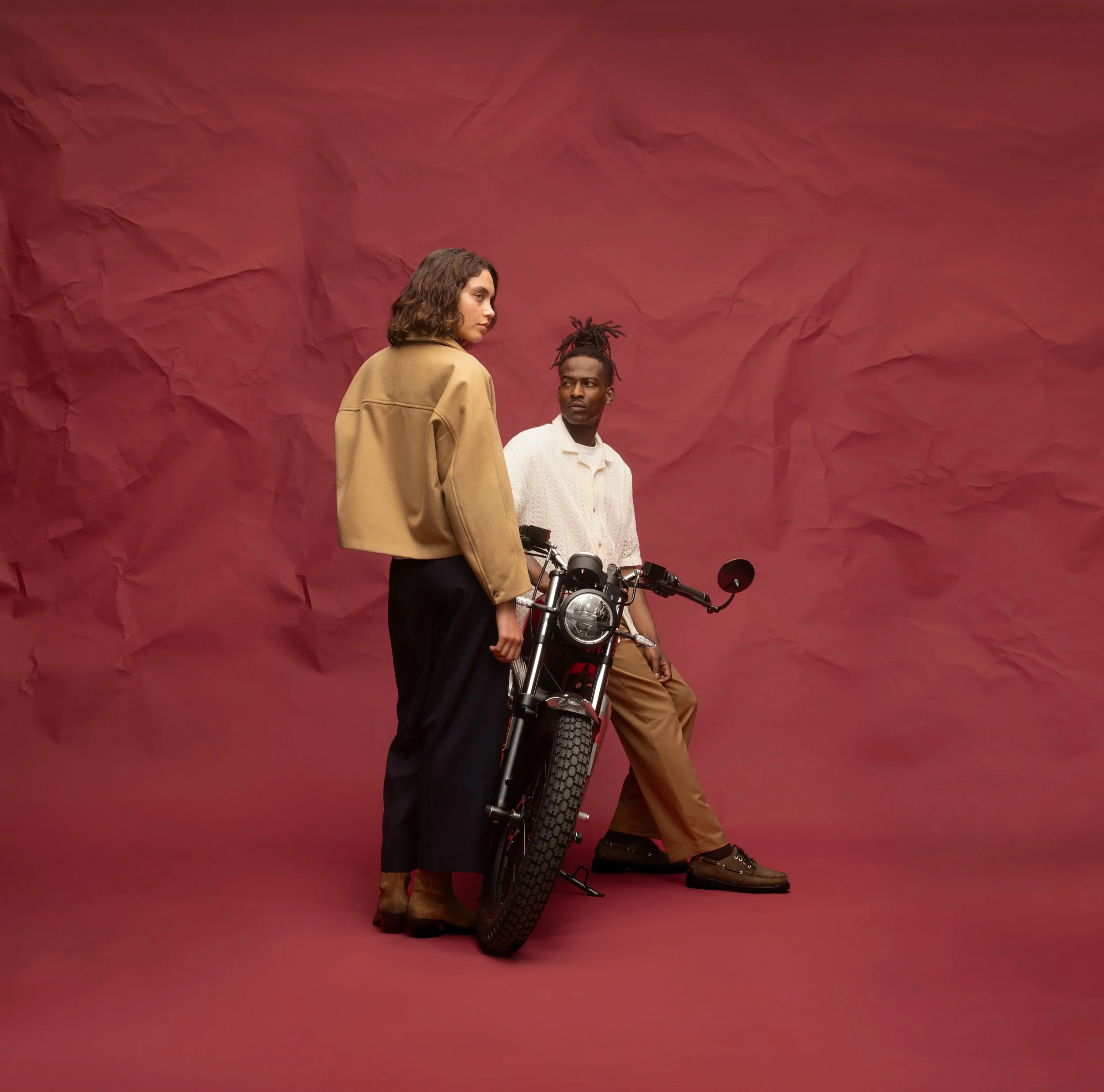 Deux personnes debout à côté d'une moto électrique contre un fond en papier brossé rouge. La femme porte un manteau beige, et l'homme porte un pull crème, portant des dreadlocks, regardant en direction de la caméra.