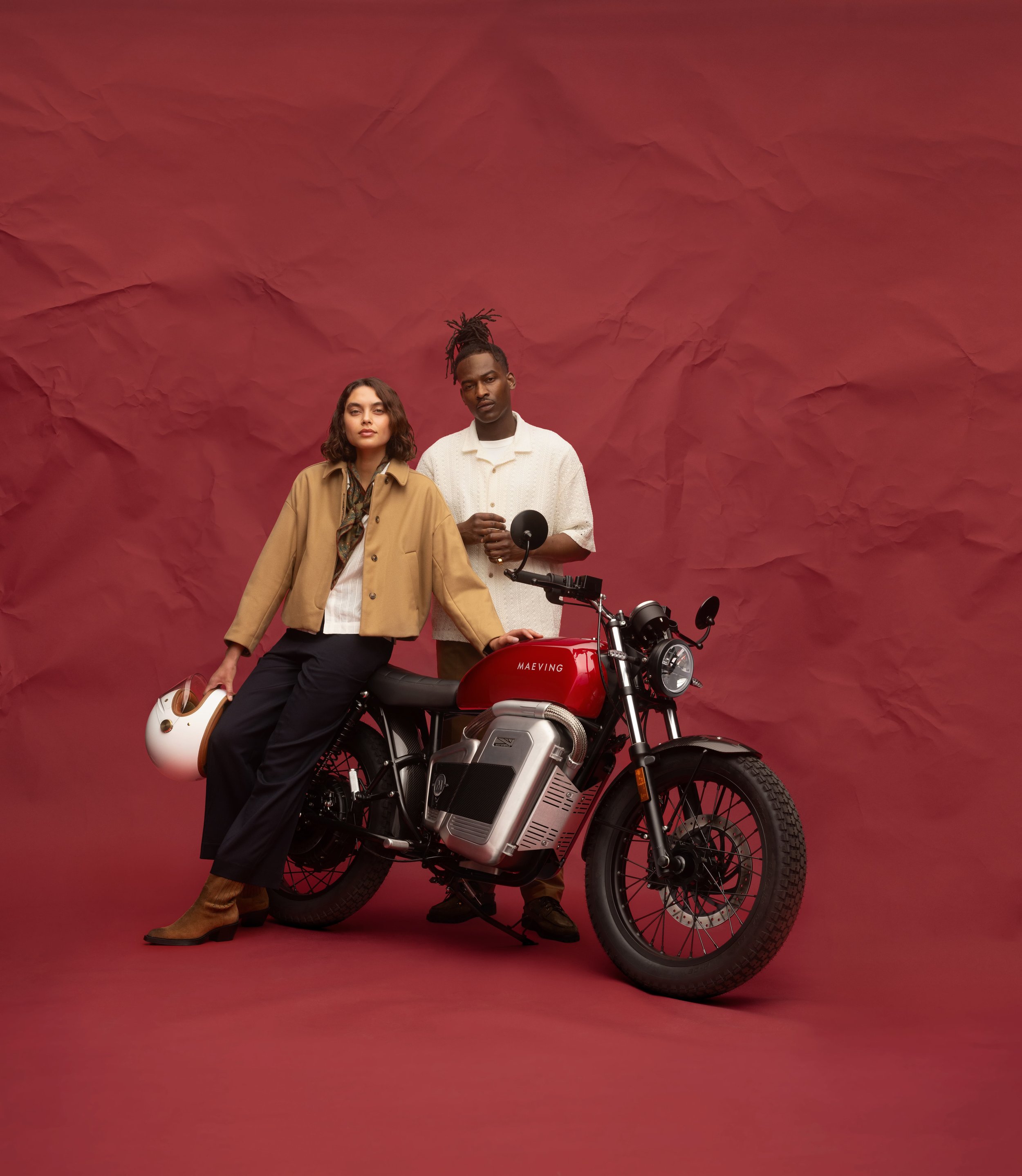 Une femme et un homme posent à côté d'une moto électrique de couleur rouge, avec un fond en papier rouge froissé.