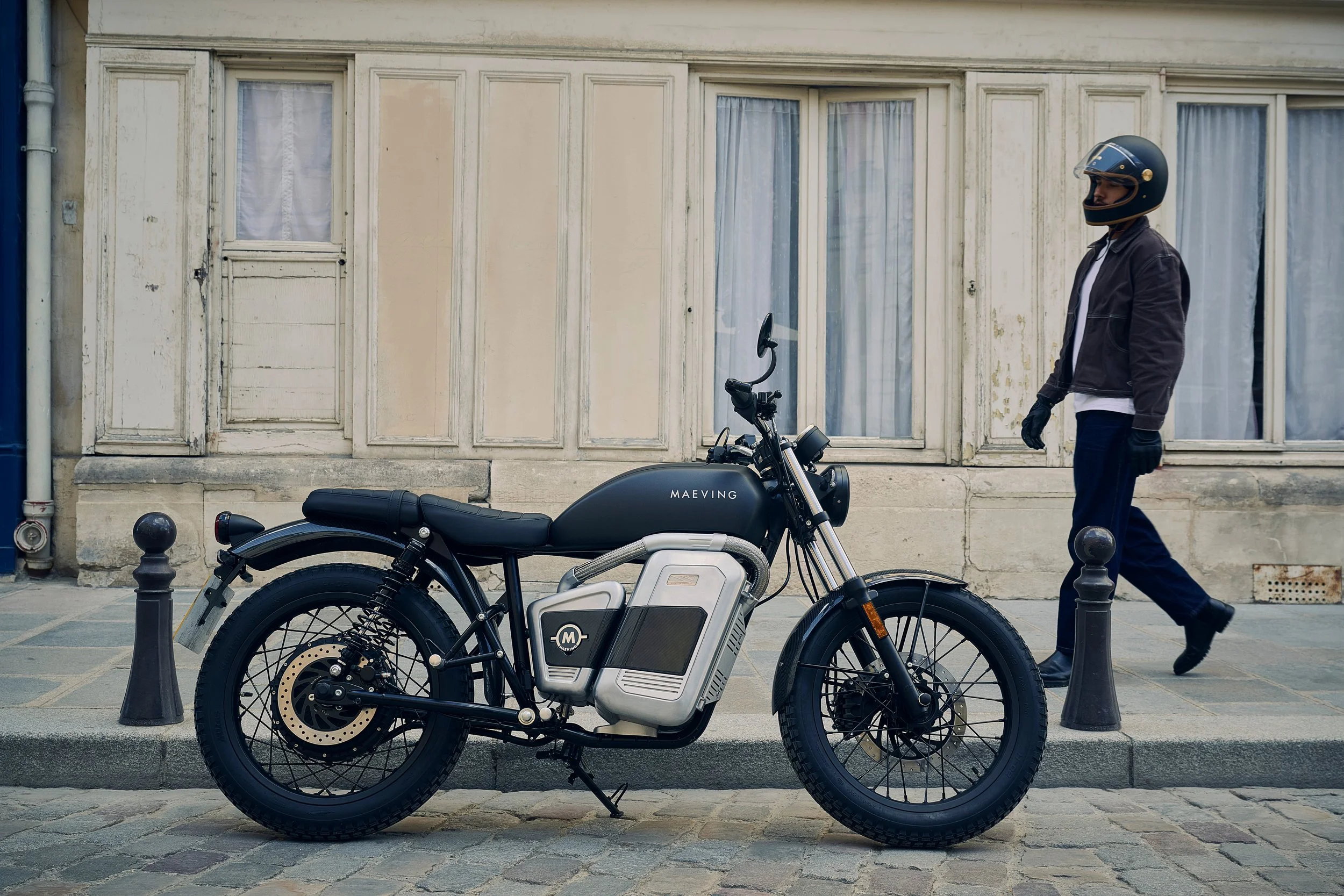 Une moto électrique noire avec le mot 'MAEVING' sur le réservoir, stationnée sur un trottoir en pierre. À l'arrière-plan, un homme portant un casque intégral et une veste sombre marche dans la rue.