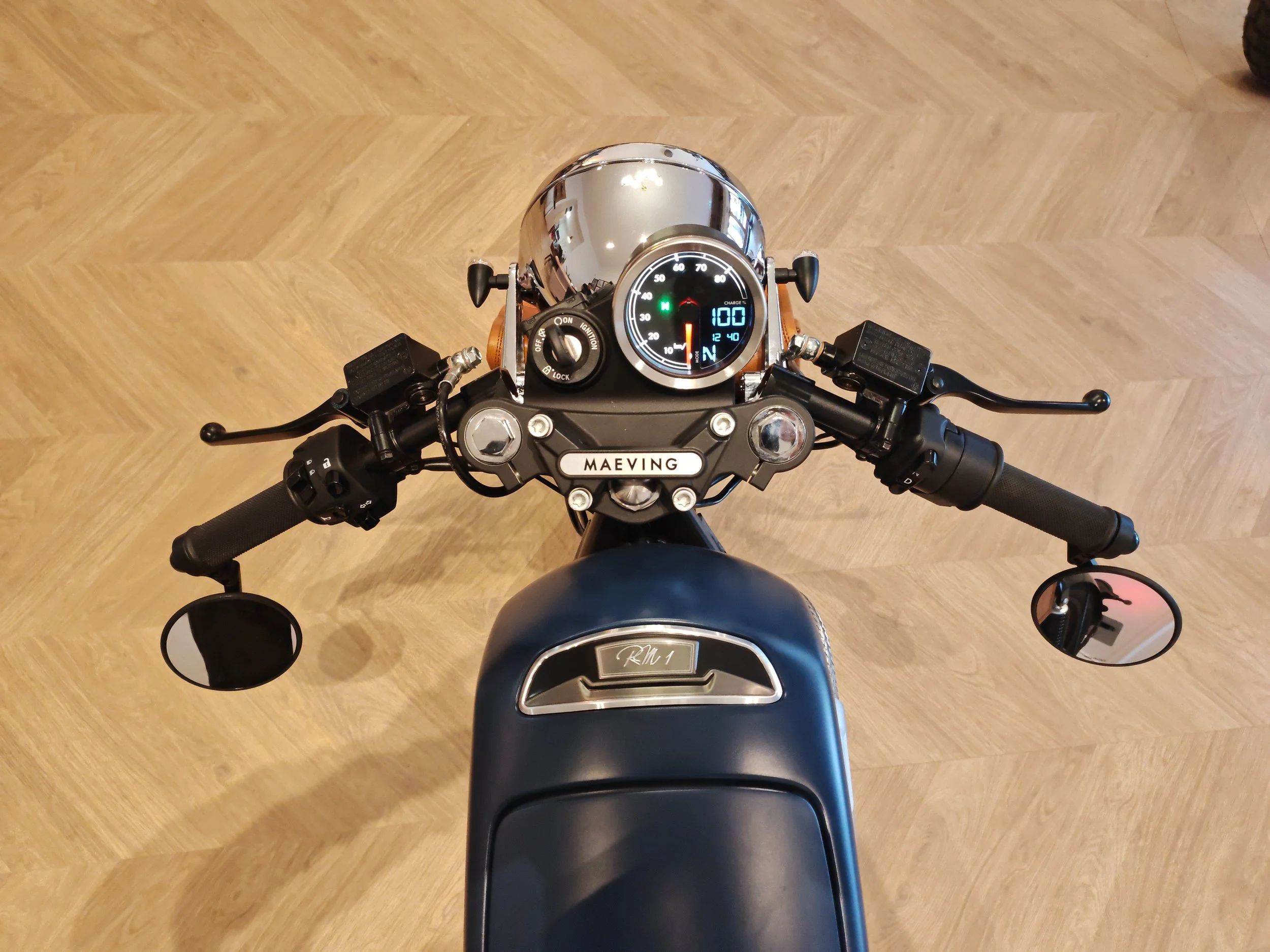 Vue du guidon d'une moto électrique rétro avec tableau de bord numérique, miroirs, et poignée, sur un sol en bois. Maeving RM1 Lyon