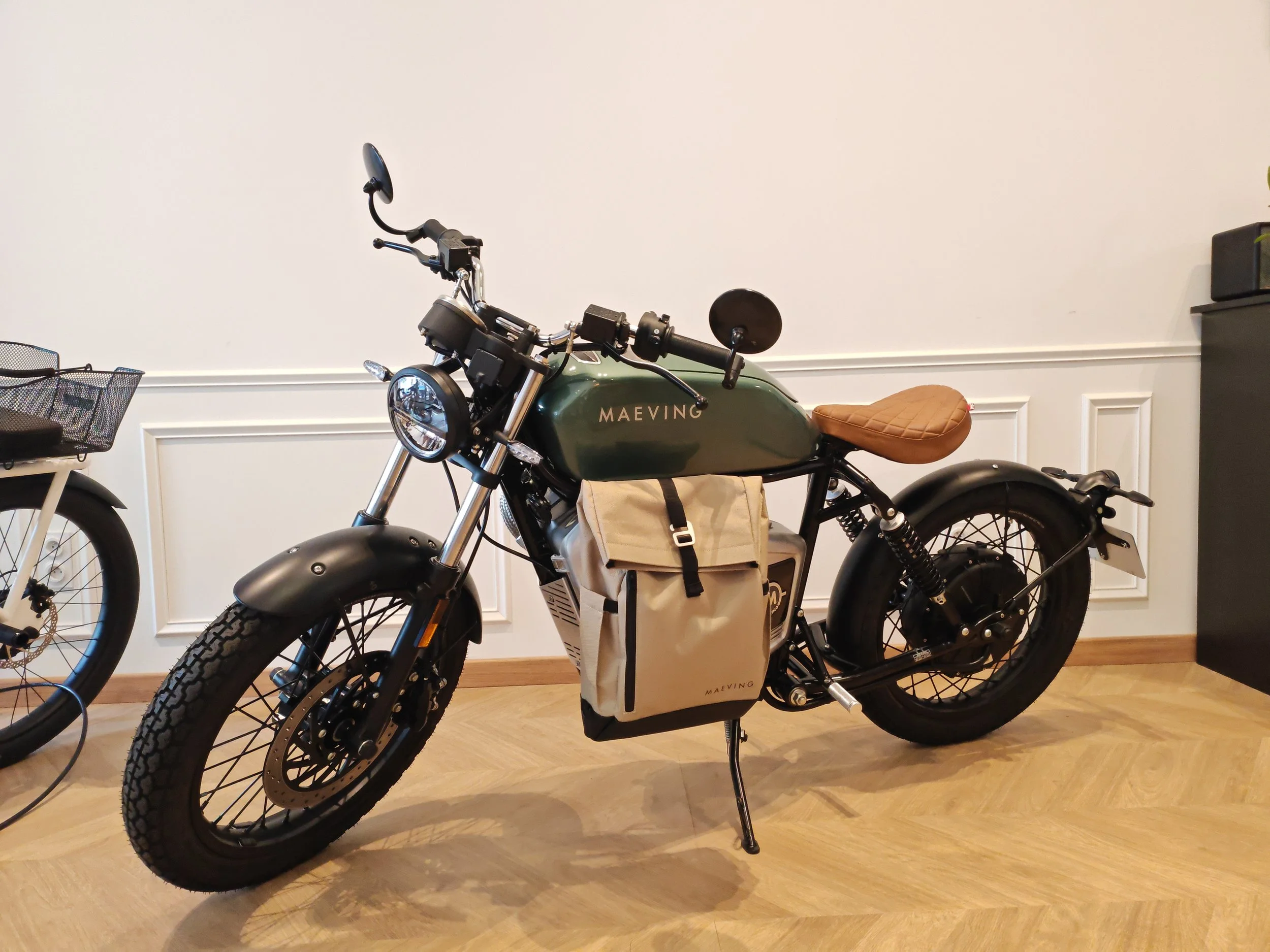 Une moto électrique verte avec un siège en cuir marron, un sac beige attaché au cadre, sur un sol en bois dans une pièce avec murs blanc. Maeving RM1S occasion Lyon.