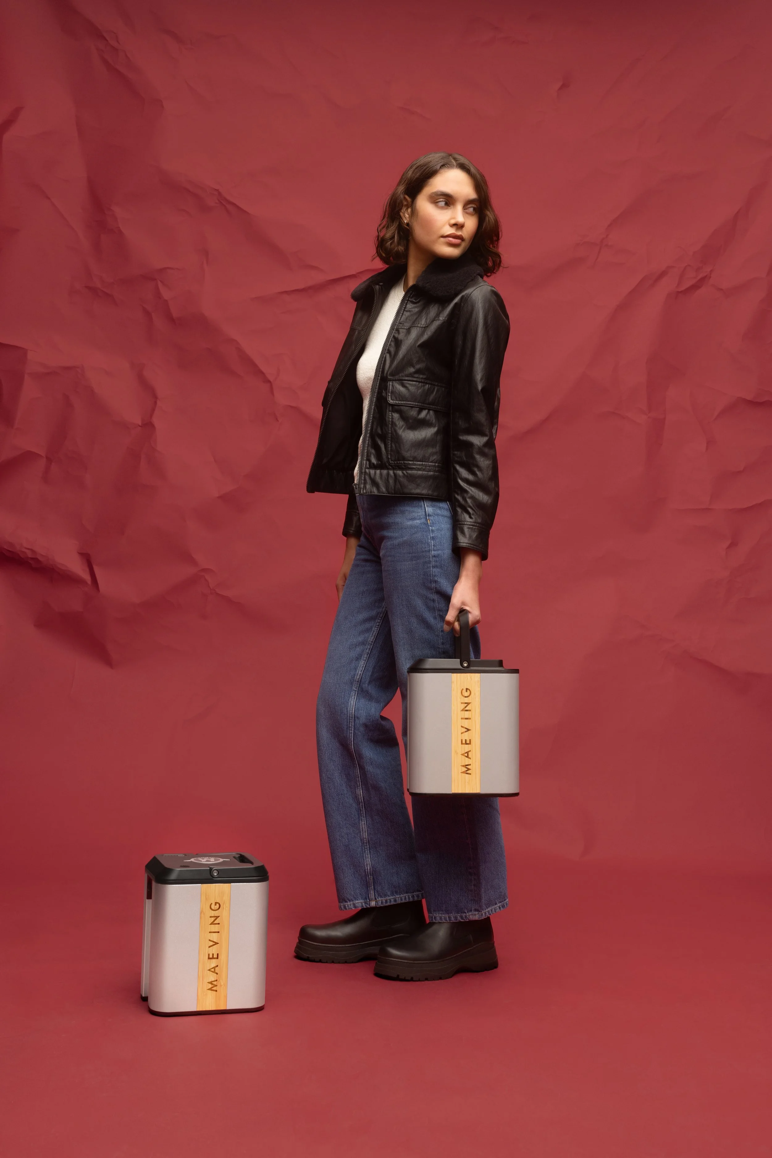 Une femme avec des cheveux bruns ondulés porte une veste en cuir noire, un pull beige, un jean et des bottes noires, tenant une batterie de moto électrique.