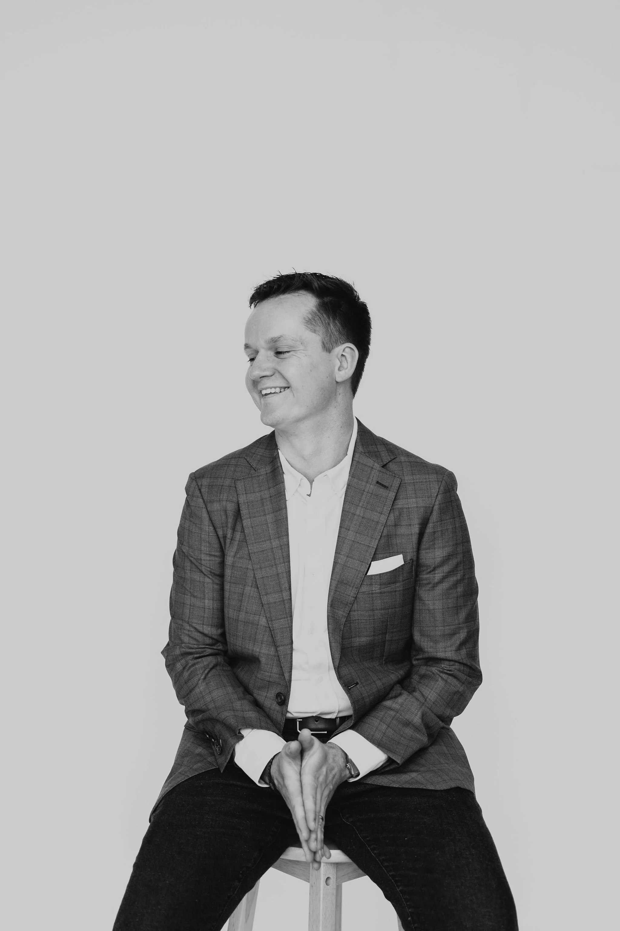 A man in a blazer and white shirt sitting on a stool, smiling, in a black and white photo.