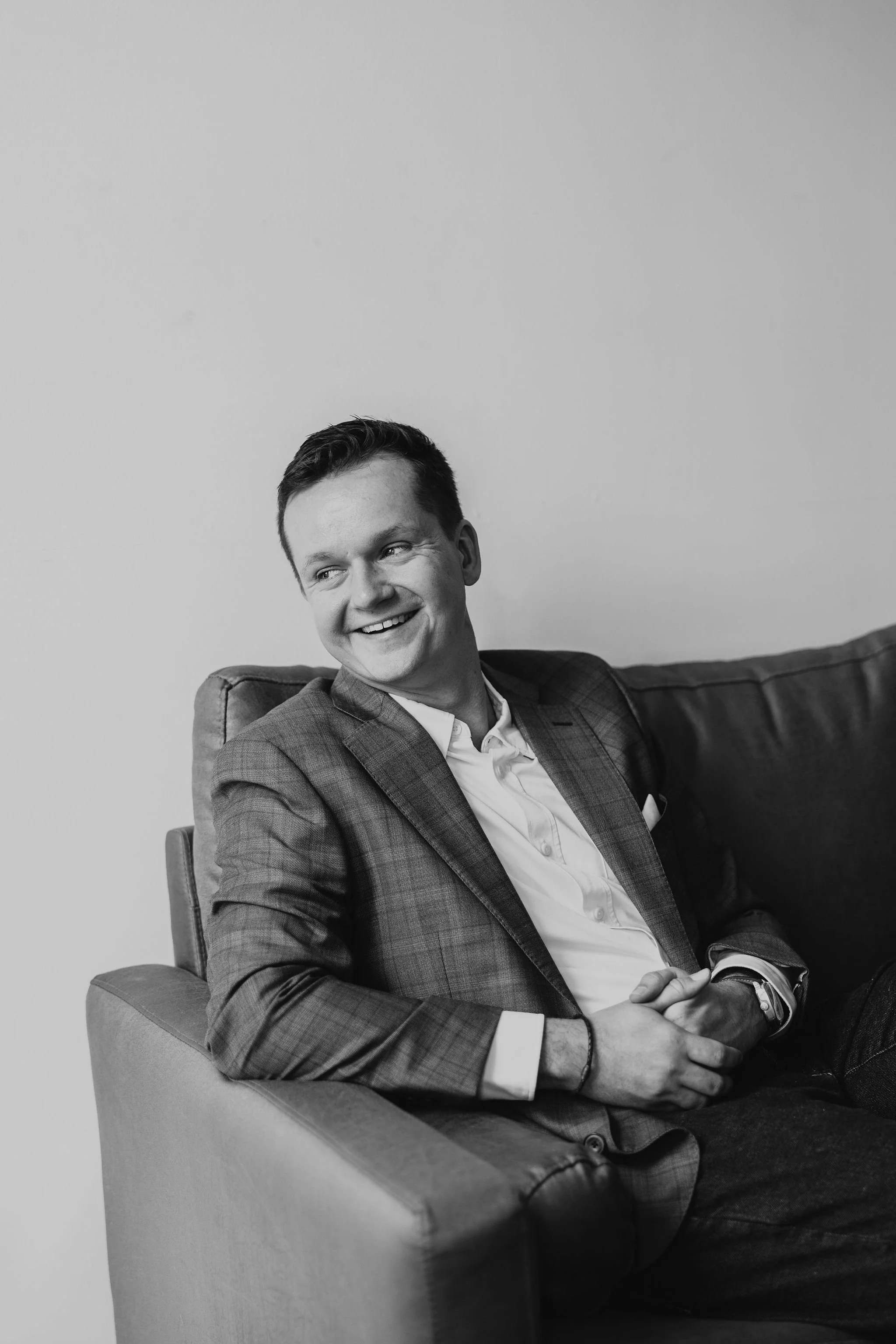 A young man in a suit sitting on a sofa, smiling and looking to his left, in a black and white photograph.